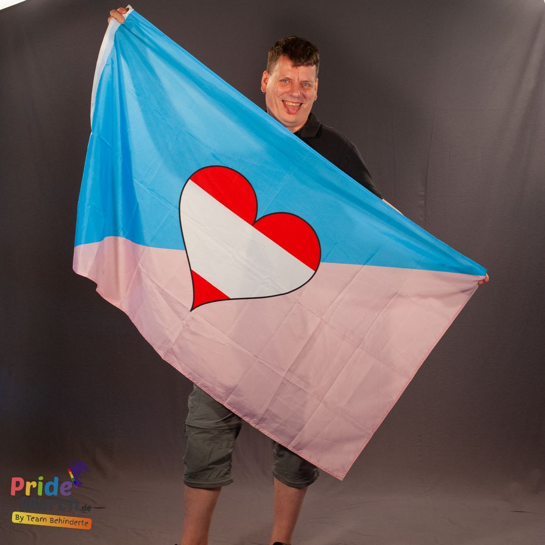 In der originalen Studioaufnahme präsentiert das ehrenamtliche Laienmodel Rene gut gelaunt die weit geöffnete Ageplay Pride Flagge vor einem dunkelgrauen Hintergrund. Er hält die große Fahne im Format 150x90 cm mit beiden Händen ausgebreitet, sodass das diagonale Farbdesign in Hellblau und Rosa mit dem zentralen rot-weiß-roten Herz-Symbol perfekt sichtbar wird. Unten links im Bild ist das Logo von PrideMerch.de (By Team Behinderte) eingebunden.