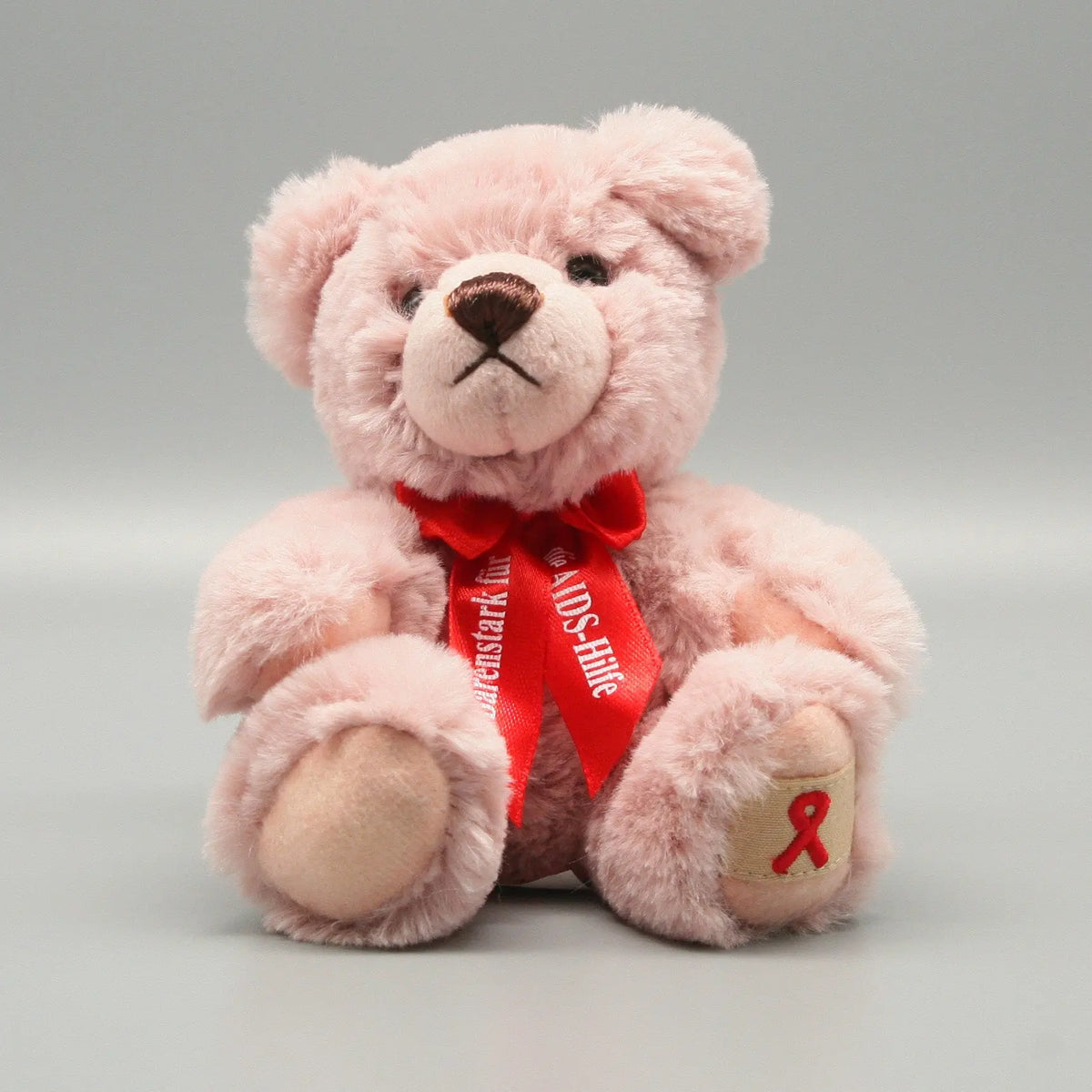 Der rosa Aids-Teddy aus dem Jahre 2023 sitzt frontal und aufrecht. Das zartrosa Plüschfell wirkt besonders weich und wird durch die markante rote Halsschleife durchbrochen. Wie bei vielen Sammlerstücken ist auch hier die rote Solidaritätsschleife auf der Fußsohle deutlich als Stickerei erkennbar.