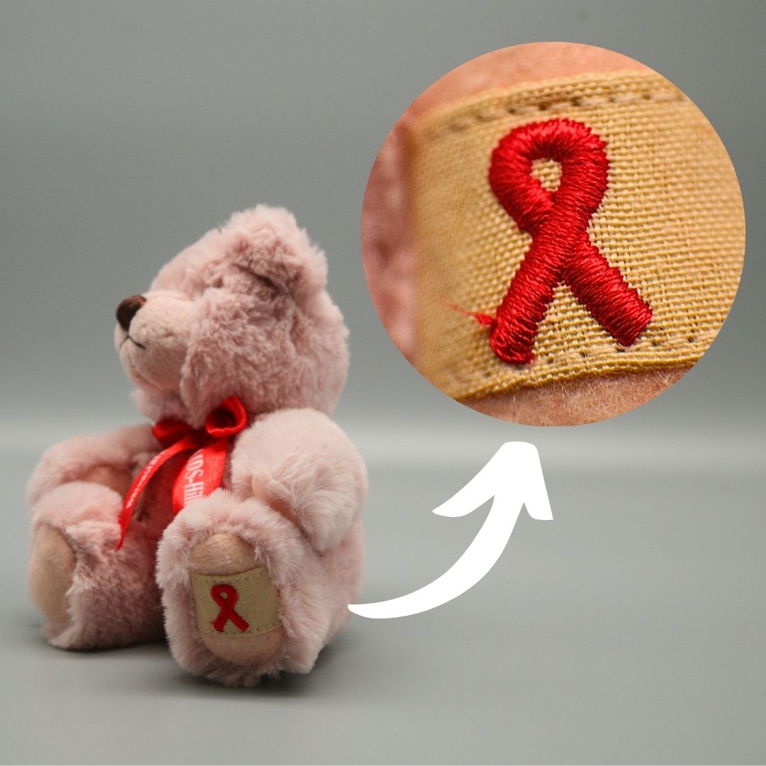 Rosafarbener AIDS-Teddy mit roter Halsschleife, seitlich sitzend auf grauem Hintergrund. Rechts vergrößert ein runder Ausschnitt das gestickte rote Red Ribbon auf der beigefarbenen Pfote.