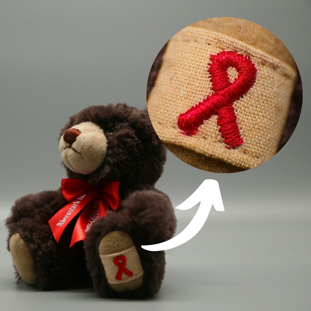Dunkelbrauner AIDS-Teddy (2021/2022) mit roter Halsschleife. Eine runde Vergrößerung zeigt die auf eine beige Pfote gestickte rote Solidaritätsschleife; ein weißer Pfeil verbindet Detail und Teddy.
