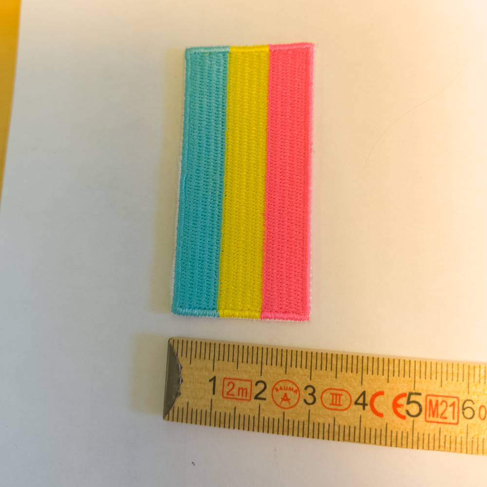 Direkt über einer gelben Zentimetermaß-Skala liegt ein schmalerer Patch mit sauberer weißer Umrandung. Die dicht gestickte Fläche zeigt das Pansexuell-Motiv mit drei gleich breiten, längs angeordneten Balken in leuchtendem Türkisblau, Gelb und Pink. Der Zollstock verrät, dass der Aufnäher etwa 3,5 cm in der Breite misst.