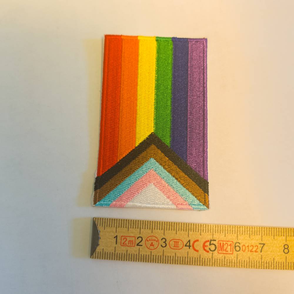 Die bekannte Progress Pride Flagge als hochwertiger Stickaufnäher, hier im Hochformat platziert. Im unteren Bereich ragt der Pfeil in Schwarz, Braun sowie den Trans-Farben nach oben in die vertikal verlaufenden Regenbogenstreifen hinein. Ein darunterliegendes gelbes Maßband zeigt, dass dieses umkettelte Modell rund fünf Zentimeter breit ist.