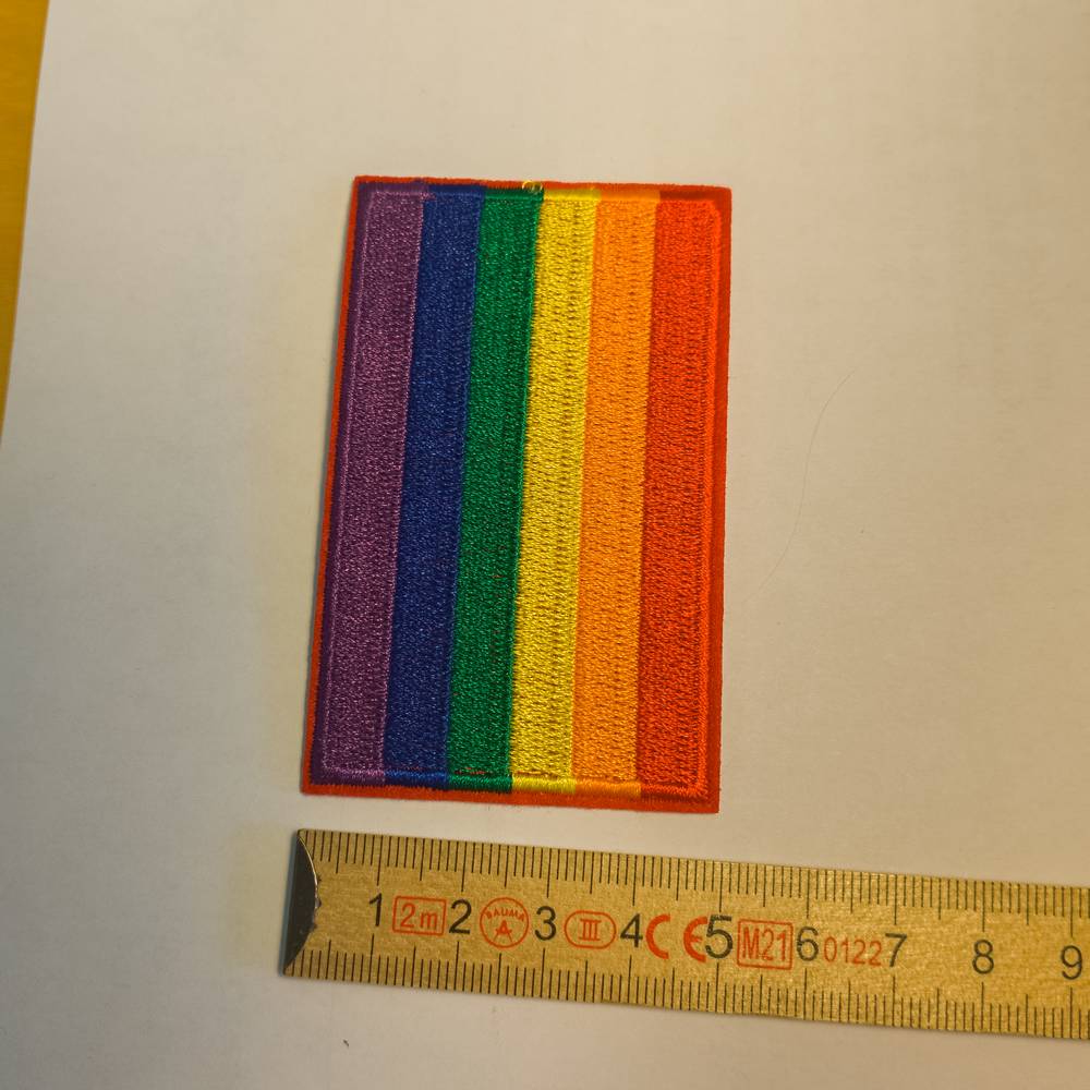 Deutlich breiter präsentiert sich dieser Regenbogen-Aufnäher. Die sechs klassischen Pride-Farben – Violett, Blau, Grün, Gelb, Orange und Rot – sind hochkant gestickt und werden von einer kräftig roten Naht komplett umrandet. Am unteren Bildrand verdeutlicht ein angelegter Gliedermaßstab eine Breite von etwa fünf Zentimetern.