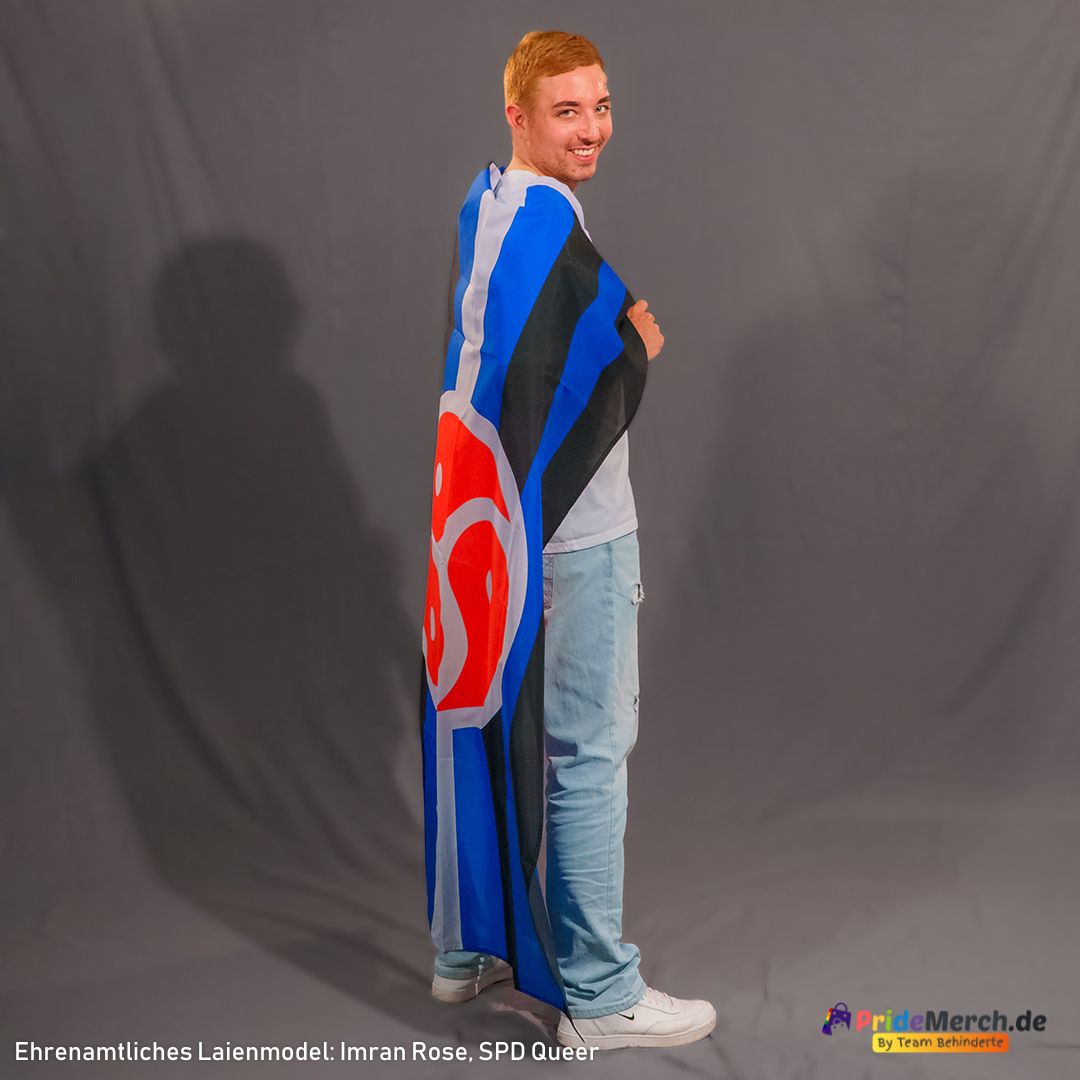Das ehrenamtliche Laienmodell Imran Rose steht lächelnd im Profil vor einem grauen Studiohintergrund und blickt über die Schulter in die Kamera. Er hat kurzes, kupferblond gefärbtes Haar, trägt ein einfaches weißes T-Shirt, eine hellblaue Jeans und weiße Sneaker. Die große BDSM-Flagge liegt wie ein langer Umhang über seinem Rücken, sodass die schwarz-blau-weißen Streifen und das kräftig rote Triskelion faltenwerfend bis zu seinen Waden herabhängen.
