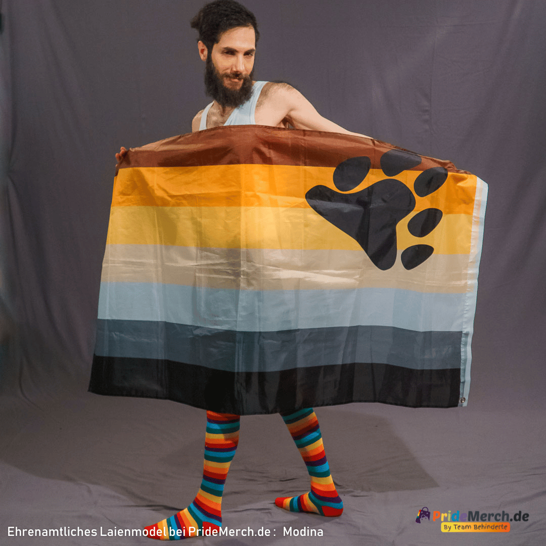 Das Laienmodell Modina steht vor einem grauen Studiohintergrund. Er hat einen dunklen Vollbart, volles dunkles Haar und trägt ein helles Tanktop sowie kniehohe, leuchtende Regenbogensocken, die einen starken Kontrast bilden. Er hält die große, erdtonfarbene Bear Pride Flagge mit beiden Händen so vor seinen Beinen und dem Unterkörper ausgebreitet, dass die große schwarze Bärentatze auf der rechten Seite gut sichtbar ist. Er blickt leicht schmunzelnd in die Kamera.
