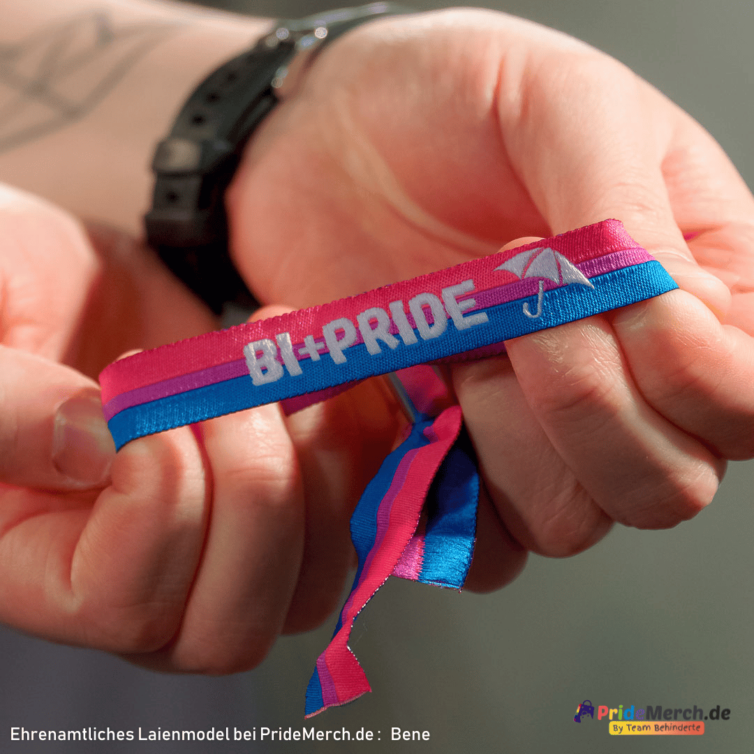 Nahaufnahme des ehrenamtlichen Laienmodels Bene, der lächelnd in die Kamera blickt und stolz seinen Arm präsentiert. Am Handgelenk trägt er das blau-pink-lilafarbene Bi+ Pride Armbändchen mit weißer „BI+PRIDE“ Stickerei und Schirm-Symbol. Unten links steht der Text „Ehrenamtliches Laienmodel: Bene“, unten rechts ist das offizielle Logo des inklusiven Online-Shops PrideMerch.de integriert.