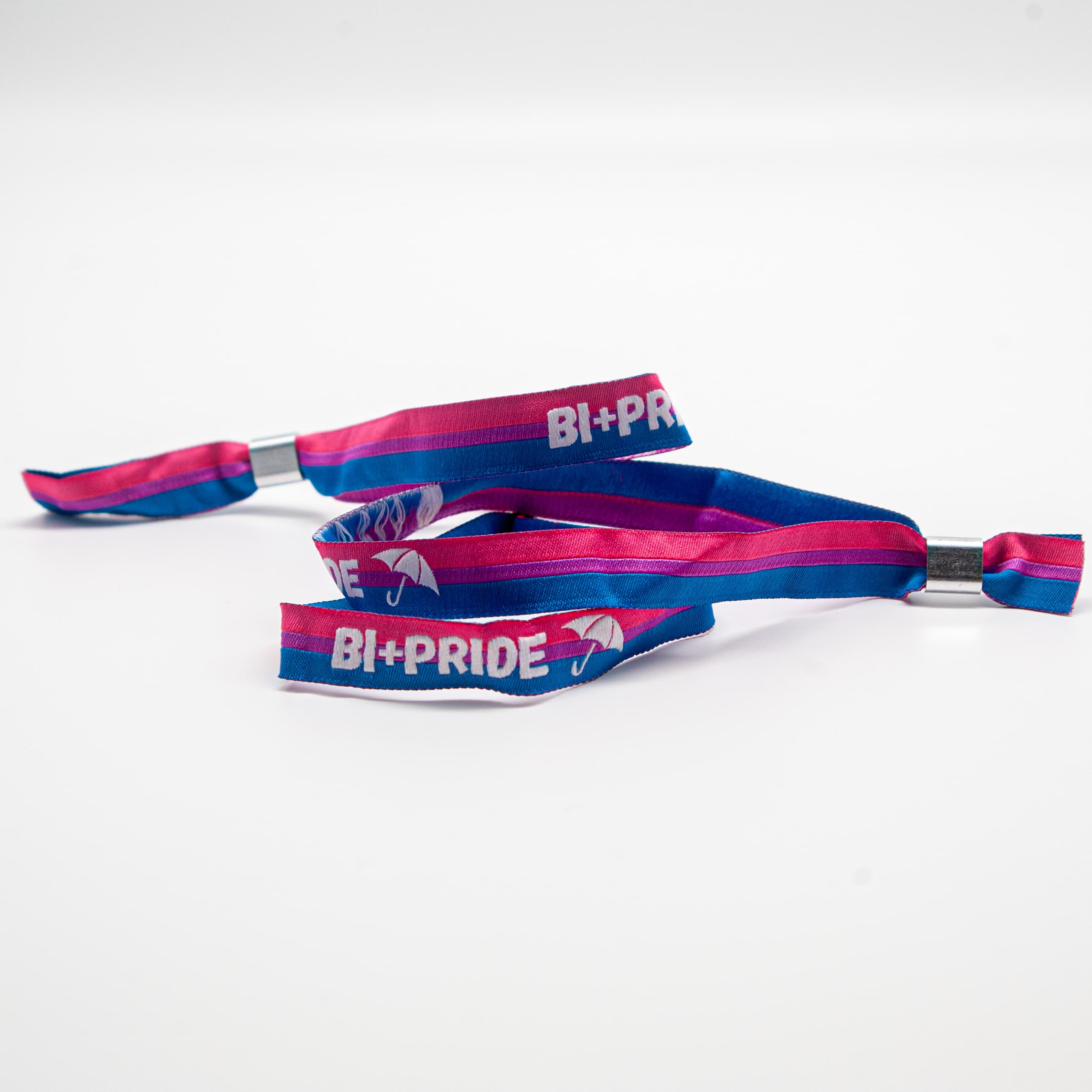 Ein gewebtes Stoffarmband in den Bi-Pride-Farben Pink, Lila und Blau liegt geschwungen auf weißem Hintergrund. Auf dem Band ist in weißer Schrift 'BI+PRIDE' sowie ein kleiner weißer Regenschirm eingestickt. Dieses Solidaritätsprodukt in Kooperation mit dem Bi+ Hamburg e.V. unterstützt das Inklusionsprojekt Team Behinderte. 10% des Umsatzes fließen direkt in den Partnerverein.
