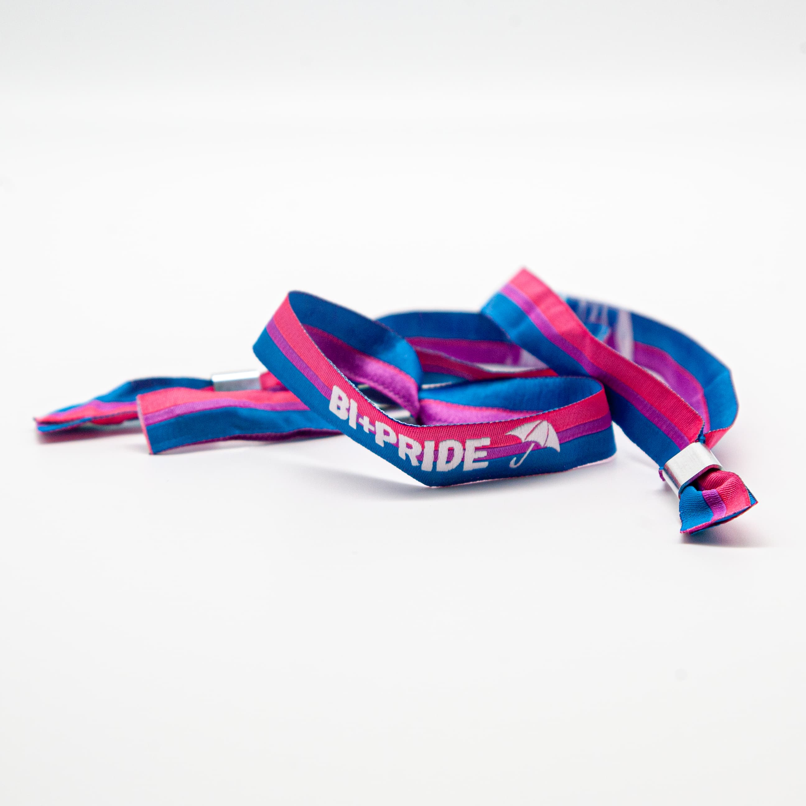 Ein flach liegendes Bi+ Pride Festivalbändchen aus weichem Stoff vor weißem Hintergrund. Die intensive Farbkombination aus Pink, Lila und Blau bringt die Pride-Stickerei „BI+PRIDE“ mit Regenschirm-Motiv perfekt zur Geltung. Zu sehen sind beide Enden des Bandes mit dem silbernen Metall-Plombenverschluss. 10 % des Umsatzes fließen direkt in die größte Bi-Demo Europas nach Hamburg.