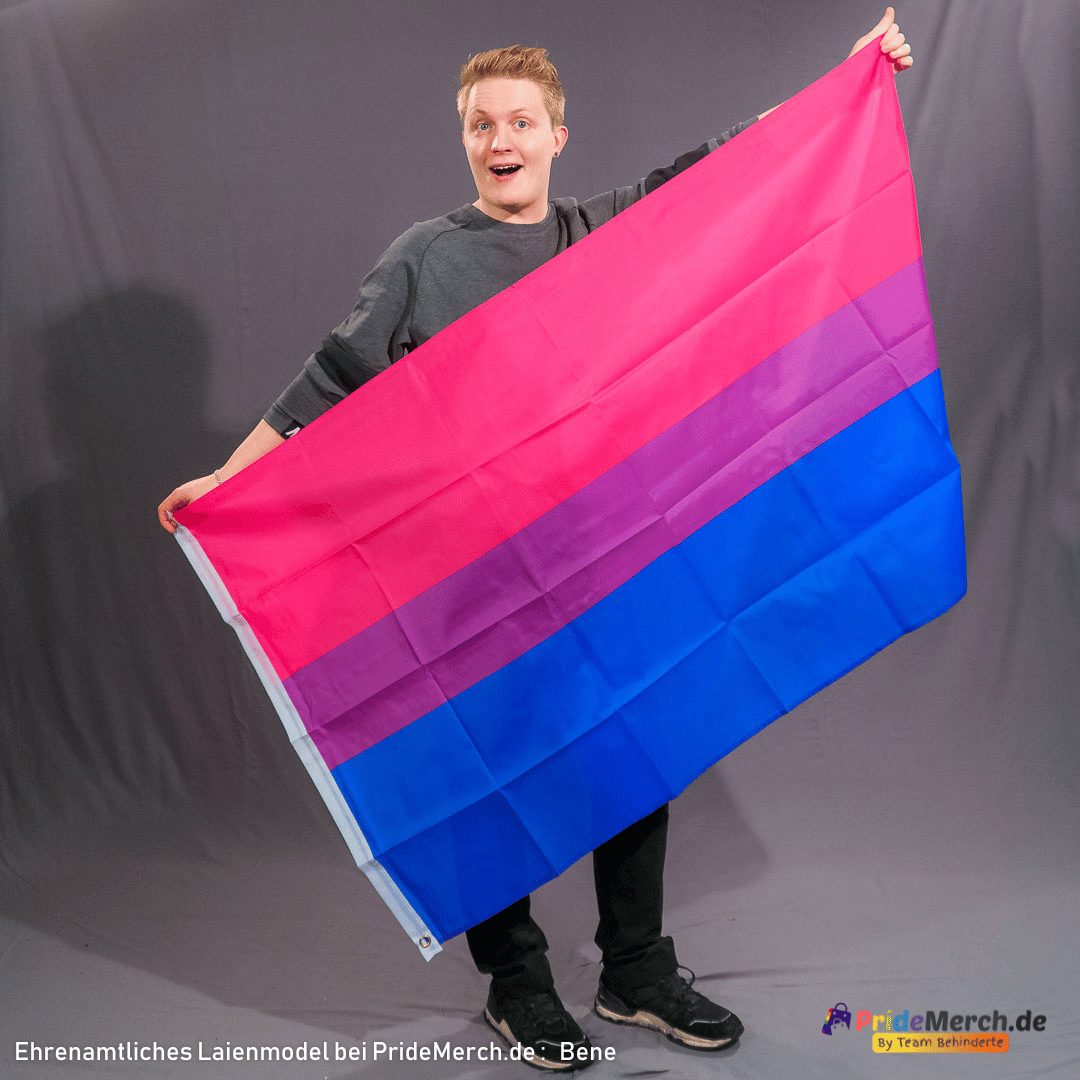 Laienmodel Bene hält die 150x90 cm große Bisexuell-Pride-Flagge mit beiden Händen weit ausgebreitet in die Kamera. Die Flagge aus wetterfestem Polyester zeigt die leuchtenden Farben der Bi+ Community in voller Pracht.