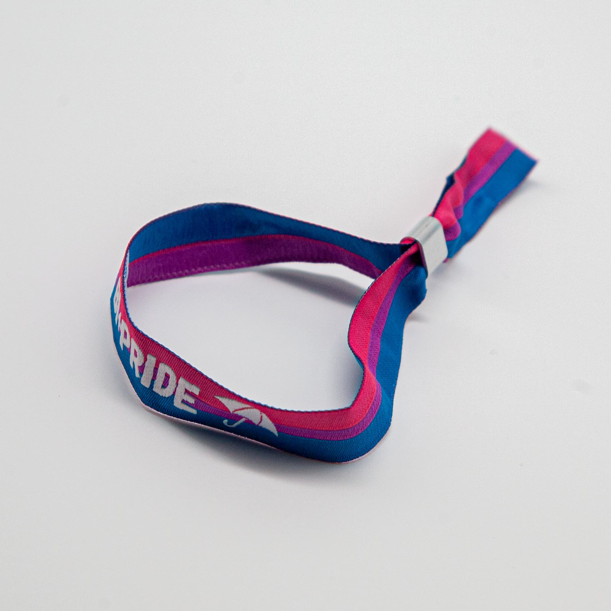 Ein weiches Stoffarmband in den horizontalen Farben der Bisexuell-Flagge (Magenta, Lavender, Royalblau) liegt geschwungen auf weißem Grund. Die weiße Einstickung zeigt das Wort „BI+PRIDE“ und das Hamburger Schirm-Logo. Dieses langlebige Accessoire für Alltag und CSD wird vom Inklusionsprojekt „Team Behinderte“ verpackt und spendet 10 % an den Bi+ Hamburg e.V.