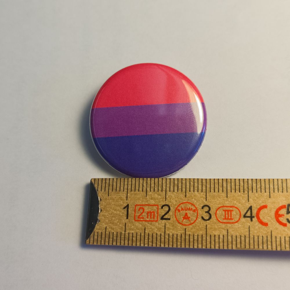 Runder Metall-Button mit den Farben der bisexuellen Flagge in Pink, Lila und Blau. Zur genauen Größendarstellung liegt der Anstecker direkt neben einem Zollstock und hat einen sichtbaren Durchmesser von 31 Millimetern.