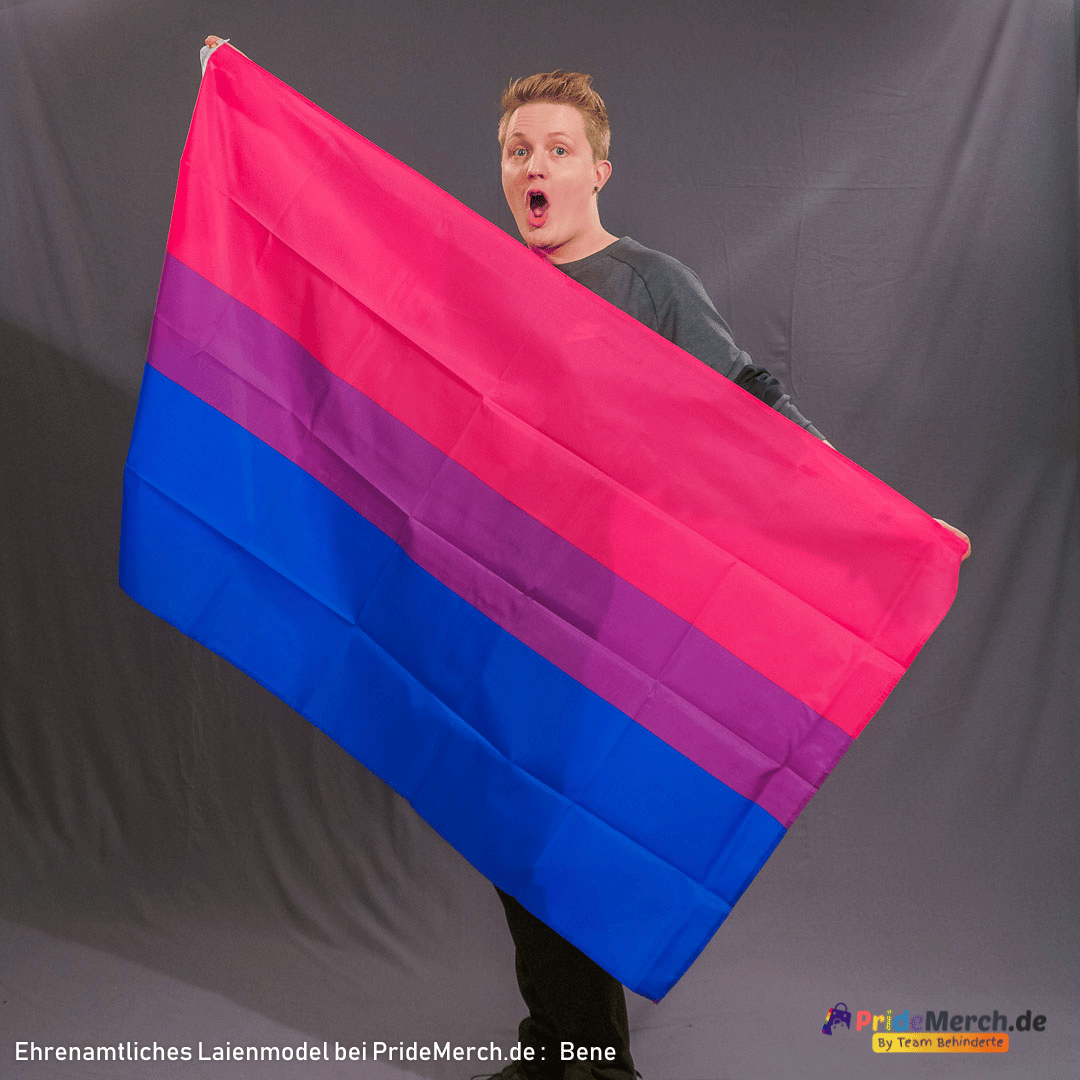Das ehrenamtliche Model Bene präsentiert die Bi-Flagge mit einem erstaunten, freudigen Gesichtsausdruck. Die Flagge wird schräg gehalten, um die Qualität des Stoffes und die Intensität der Farben Pink, Lila und Blau zu verdeutlichen.