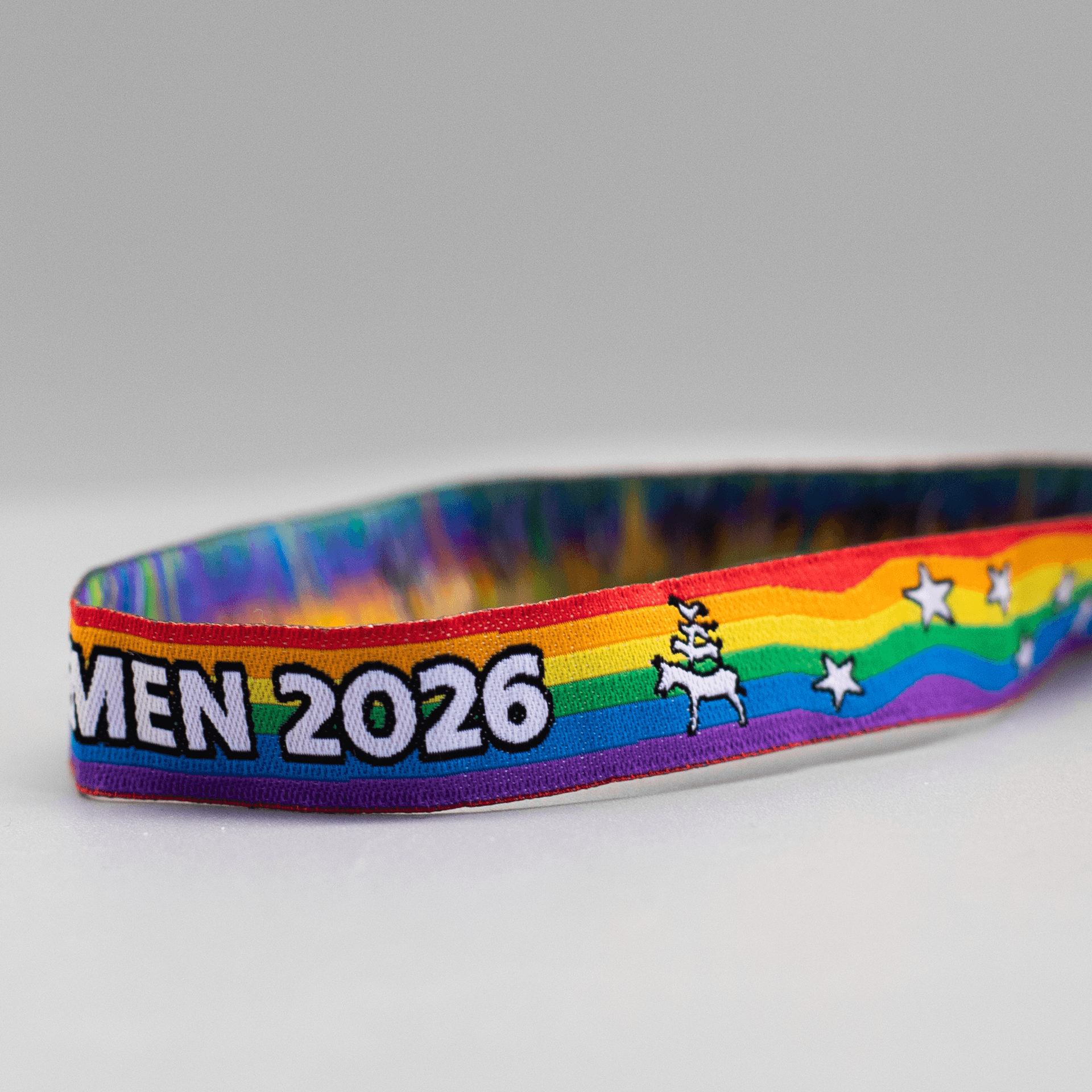 Detailaufnahme eines regenbogenfarbenen Pride-Armbändchens auf hellgrauem Hintergrund. Zu sehen ist der rechte Teil des Bandes mit dem Text „...MEN 2026“ im neuen Design: weiße Schrift mit schwarzer Umrandung. Rechts neben der Jahreszahl ist eine kleine weiße Silhouette der Bremer Stadtmusikanten mit schwarzer Kontur abgebildet. Daneben verlaufen auf den Regenbogenstreifen mehrere weiße, schwarz umrandete Sterne. Die Innenseite des Bandes zeigt ein bunt schimmerndes Muster.
