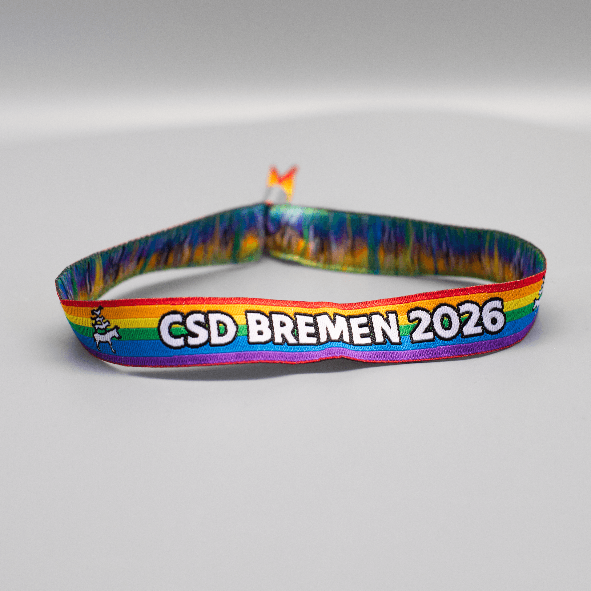 Gesamtansicht des Pride-Armbändchens für den CSD Bremen als Solidaritätsprodukt. Das Stoffband liegt als Oval auf einem hellgrauen Untergrund. Auf den klassischen Regenbogenstreifen steht zentriert der vollständige Schriftzug „CSD BREMEN 2026“ in weißer Schrift mit schwarzer Umrandung. Links neben dem Text ist als kleines Detail die Silhouette der Bremer Stadtmusikanten (weiß mit schwarzer Kontur) eingewebt.