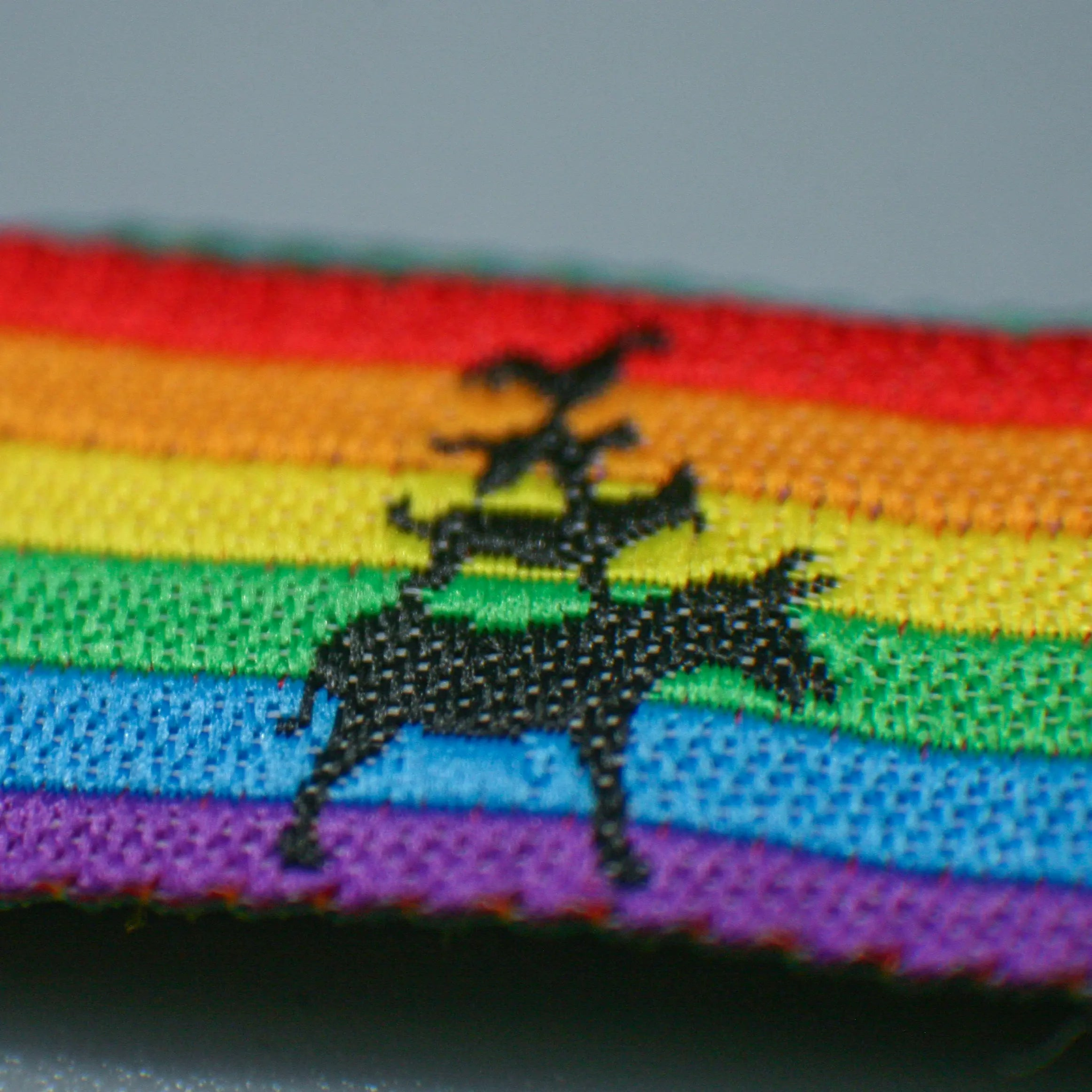Detaillierte Nahaufnahme des gewebten CSD Bremen Pridebändchens. Die schwarze Silhouette der Stadtmusikanten hebt sich hochwertig vom farbintensiven Regenbogen-Stoff ab – langlebige Qualität, die dein queeres Statement im Alltag sicher am Handgelenk hält.