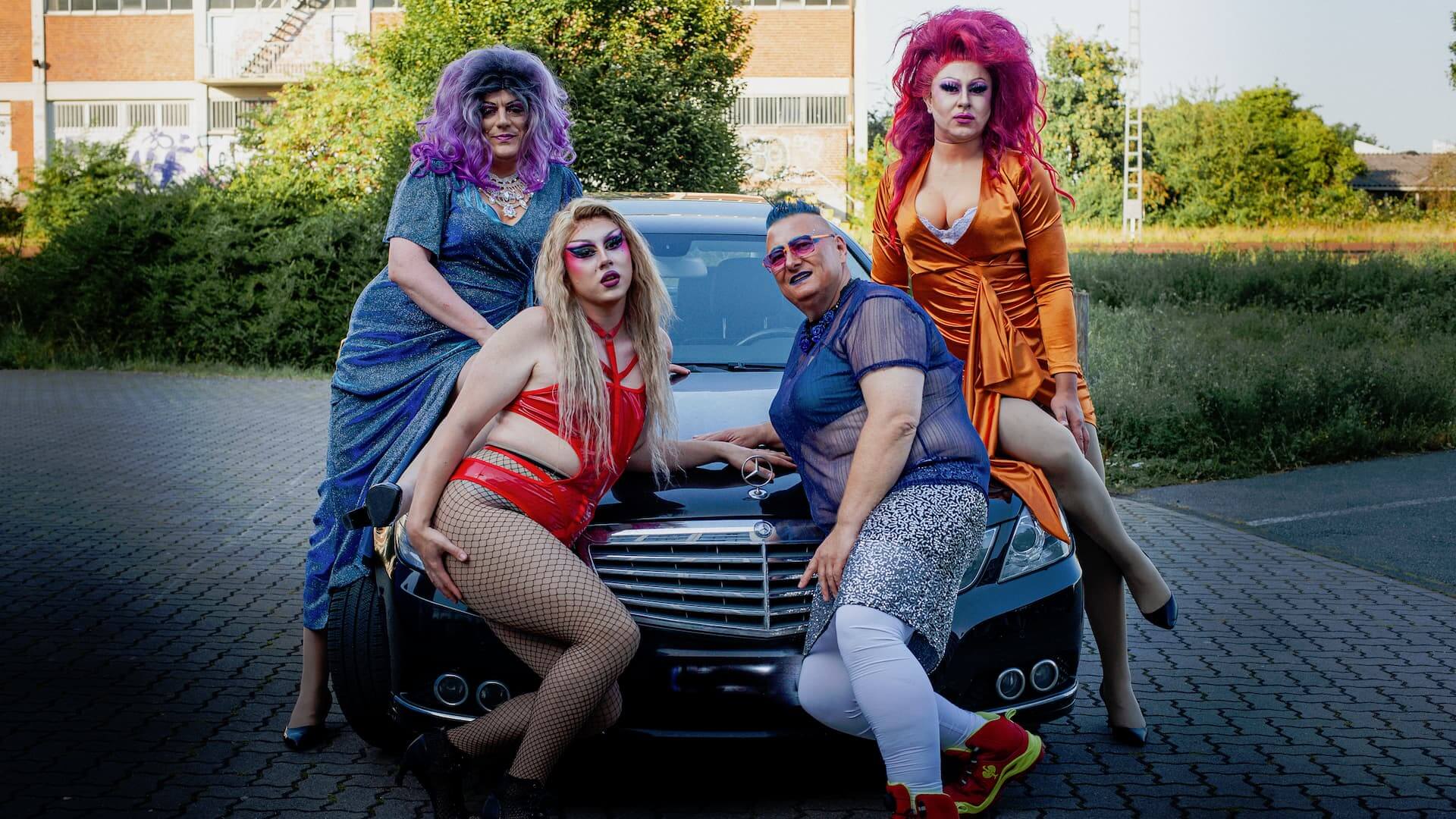 Vier Drag-Queens posieren selbstbewusst vor einer schwarzen Mercedes-Limousine auf einem gepflasterten Hof. Von links nach rechts: Jolina Wings (violette Perücke, blau-glitzerndes Kleid), Luna Mattel (blonde Haare, rotes Latex-Outfit mit Netzstrumpfhose), Gina Petrolia (blau-violettes durchsichtiges Oberteil, silberner Glitzerrock), Miss Joyce DeLone (pinke Perücke, orange Satinrobe). Hintergrund: Gebäude, Bäume und Sträucher bei sonnigem Licht.