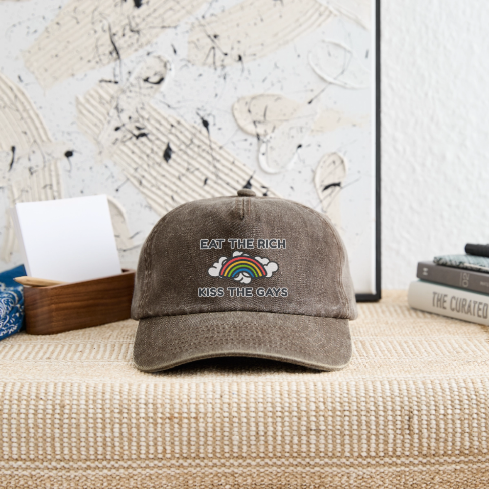 Präsentation der braunen Vintage Cap liegend in einem stilvollen Raum-Setting. Die aktivistische Doppelbotschaft „Eat The Rich Kiss The Gays“ und der leuchtende Regenbogen machen die Retro-Mütze zum perfekten Accessoire für den Straßenprotest.