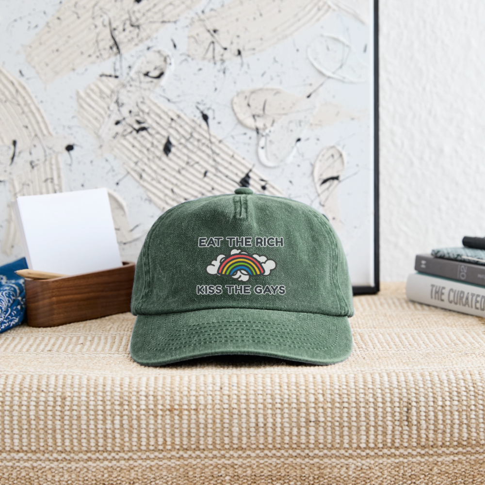 Eine flaschengrüne Vintage Cap ruht auf einer geflochtenen Ablage und rückt das Design „Eat The Rich Kiss The Gays“ in den Fokus. Luftig und provokant zugleich – das Regenbogen-Motiv zeigt selbstbewussten queeren Aktivismus.
