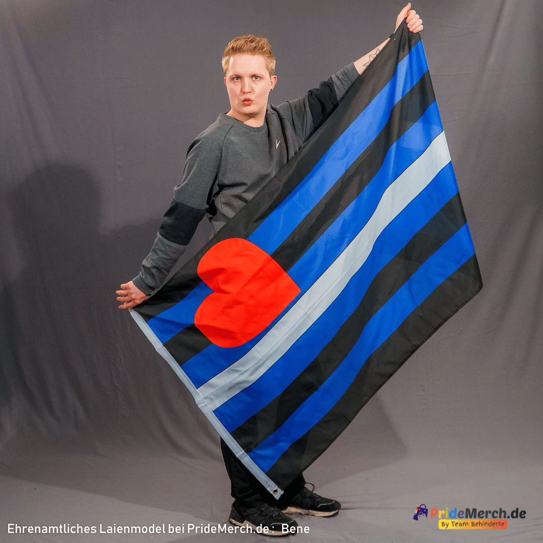 Das Laienmodell Bene blickt direkt in die Kamera und formt einen Kussmund. Er hat den linken Arm weit nach oben gestreckt und hält die Leather Pride Flagge an der oberen Ecke fest, während die rechte Hand die Fahne auf Hüfthöhe abspannt. Die schwarz-blau gestreifte Flagge mit dem weißen Mittelstreifen und dem roten Herz fällt dadurch in einer dynamischen Schräge vor seinem Körper ab.