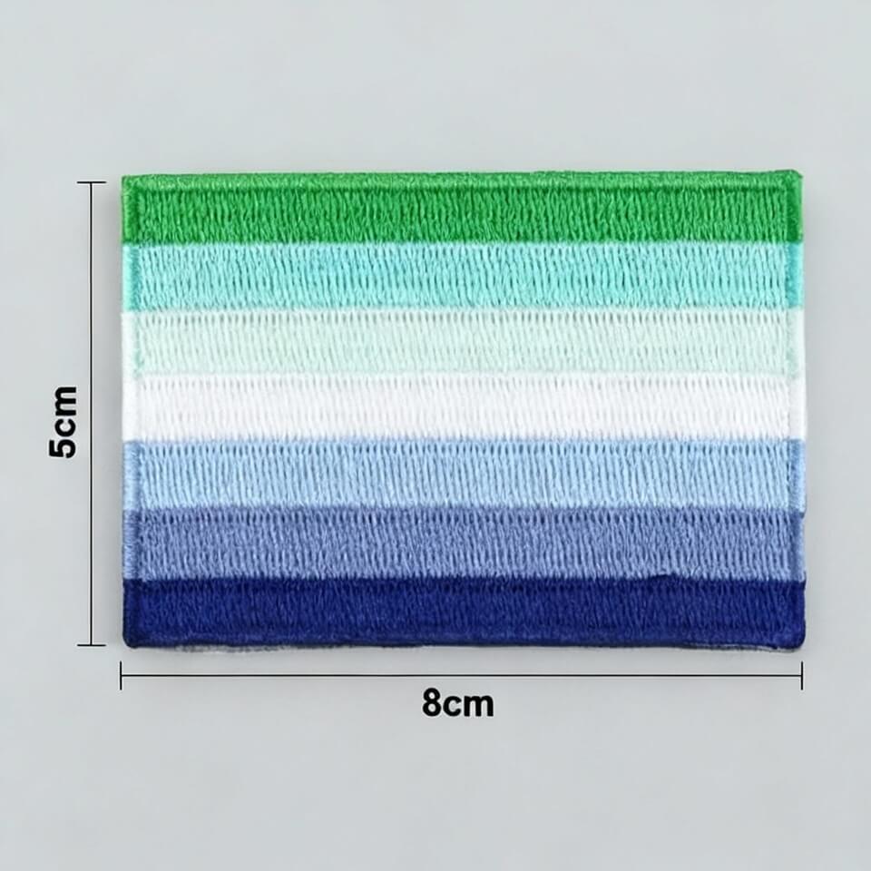 Ein dicht gestickter Bügelpatch mit den horizontalen Streifen der Gay Men (MLM) Flagge in verschiedenen Abstufungen von Grün, Weiß und Blau. Der farbintensive Aufnäher liegt auf einem hellen Hintergrund direkt an der Zentimeter-Skala eines gelben Holz-Zollstocks.