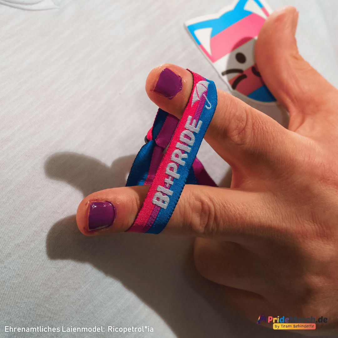 Porträt des ehrenamtlichen Laienmodels Gina Petrolia, die mit einem charmanten Lächeln für PrideMerch.de posiert. Sie blickt in die Kamera und hält ihre Hand an die Wange, wodurch das pink-lila-blaue Bi+ Pride Stoffarmbändchen an ihrem Handgelenk perfekt präsentiert wird. Unten links ist der Schriftzug „Ehrenamtliches Laienmodel: Gina Petrolia“ eingeblendet, rechts das Logo von PrideMerch.de – By Team Behinderte.