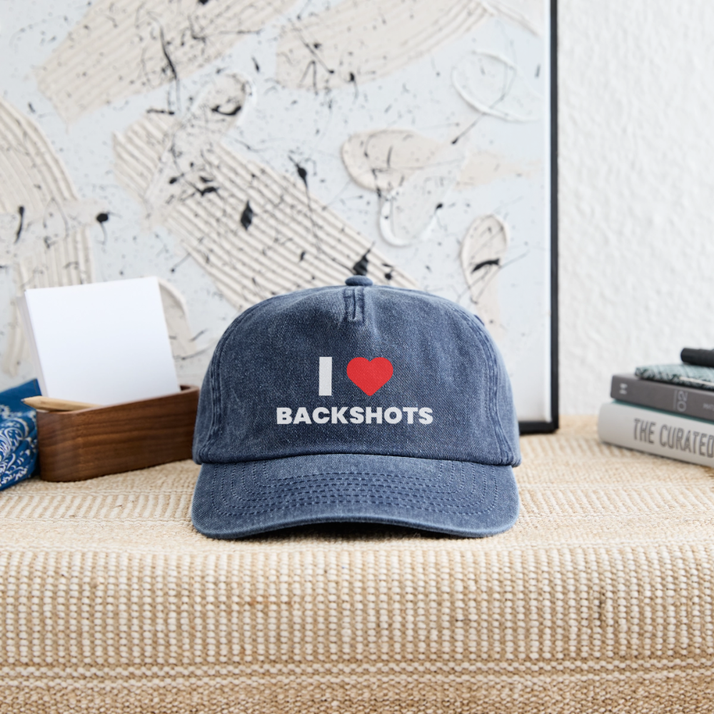 Die blaue Denim-Mütze liegt dekorativ auf einer hölzernen Kommode in der Garderobe. Der unzensierte Spruch „I ❤️ BACKSHOTS“ auf der Vorderseite kommt im warmen Licht gut zur Geltung und setzt ein humorvolles Statement.