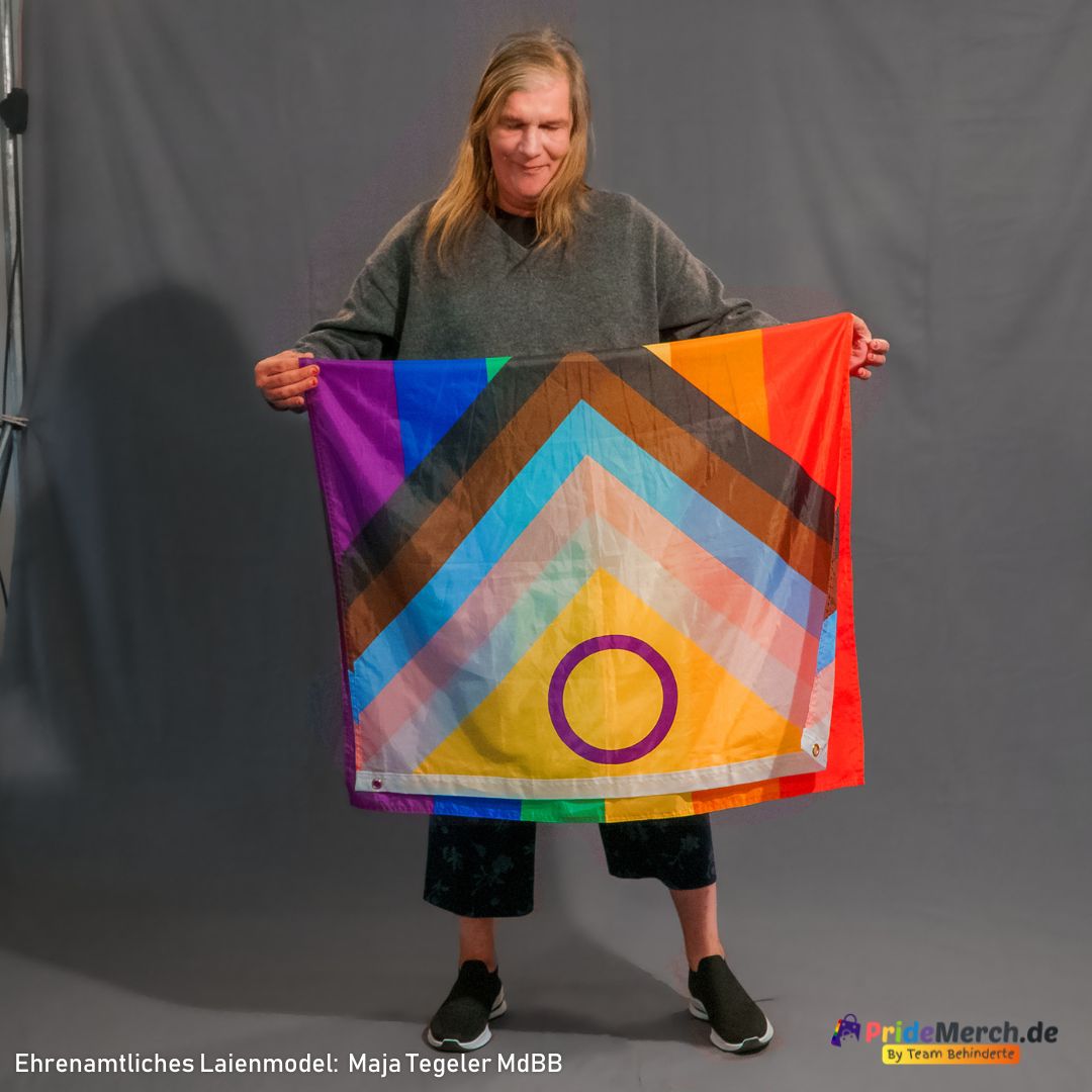 Das Laienmodell Maja Tegeler hält die erweiterte Regenbogenfahne im hellen Tageslicht. Diese Perspektive betont die feine Stoffstruktur der wetterfesten Ösenflagge sowie das harmonische Zusammenspiel der inklusiven Keil-Elemente für queeren Aktivismus.