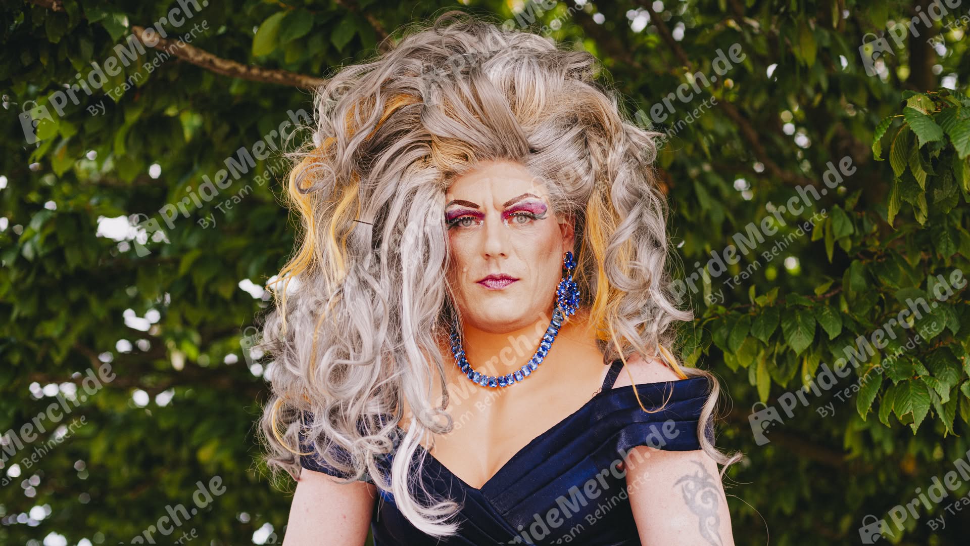 Jolina Wings in einem Drag-Look mit einer auffälligen, toupierten grau-blonden Perücke und einem eleganten blauen Outfit.