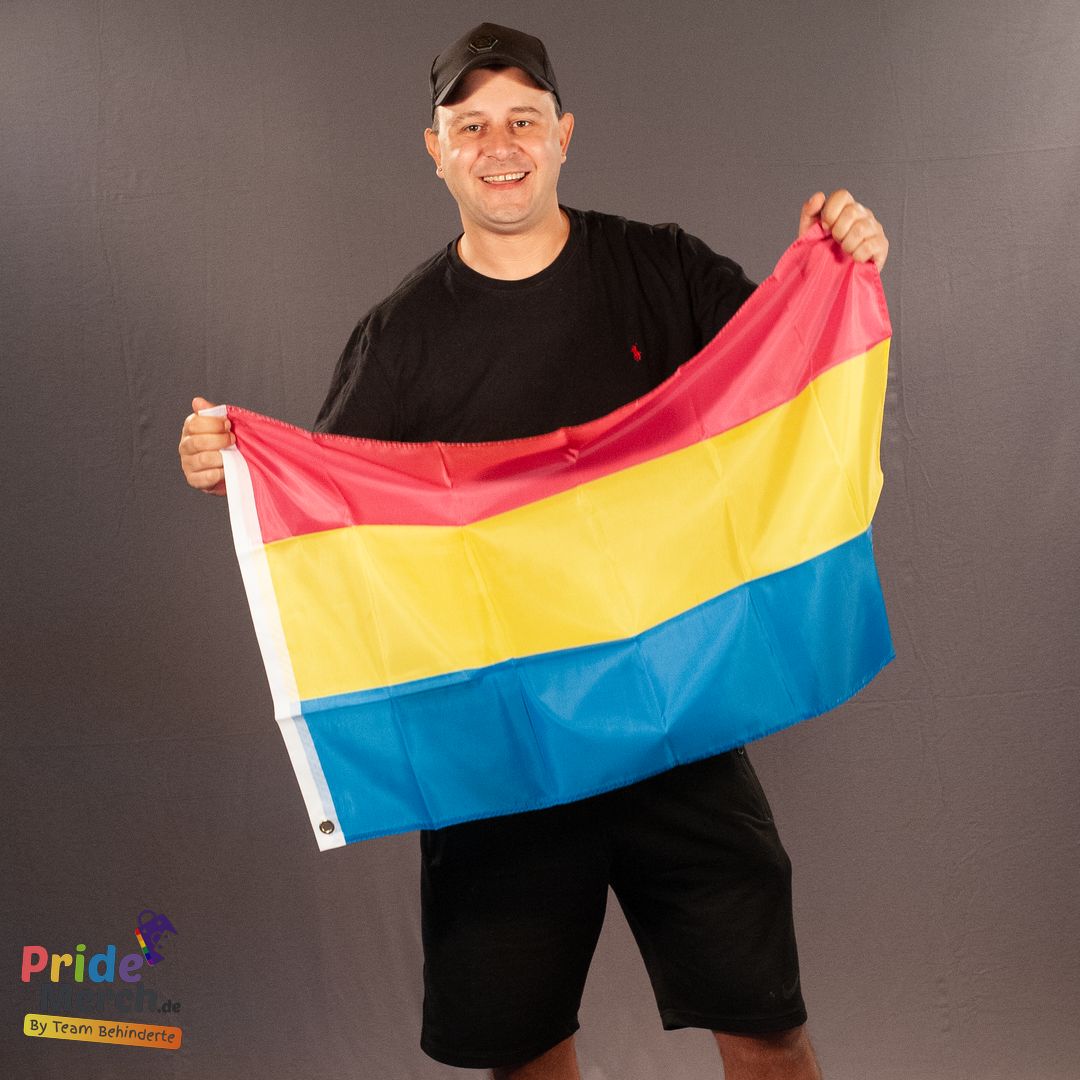 Das ehrenamtliche Laienmodell Joyce DeLone (out of Drag) steht freundlich lächelnd vor einem grauen Stoffhintergrund. Er trägt eine schwarze Basecap, ein schwarzes T-Shirt und schwarze sportliche Shorts. Mit beiden Händen hält er die kompakte, kleinere Version der Pansexuell Pride Flagge vor seinem Oberkörper. Die horizontalen Streifen in leuchtendem Pink, Gelb und Blau heben sich mit starkem Kontrast von seiner komplett schwarzen Kleidung ab. Am linken Rand der Flagge ist der weiße Saum zu sehen.
