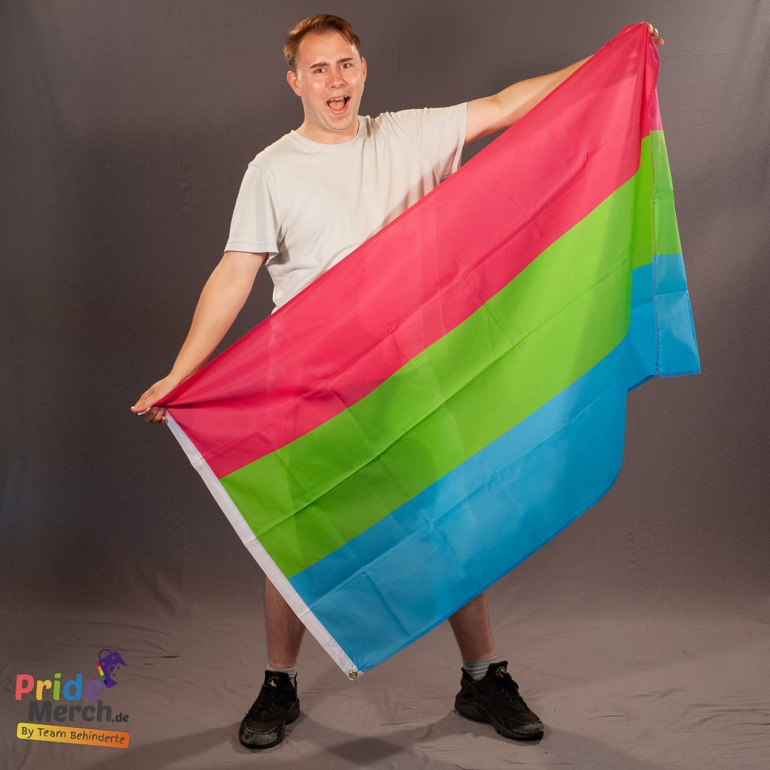 Das Bremer Laienmodell Drag Queen Luna präsentiert lachend und mit weit geöffnetem Mund die polysexuelle Pride-Fahne im Studio. Die zwei Metallösen an der weißen Kante der Flagge sind unten links im Bild gut erkennbar.