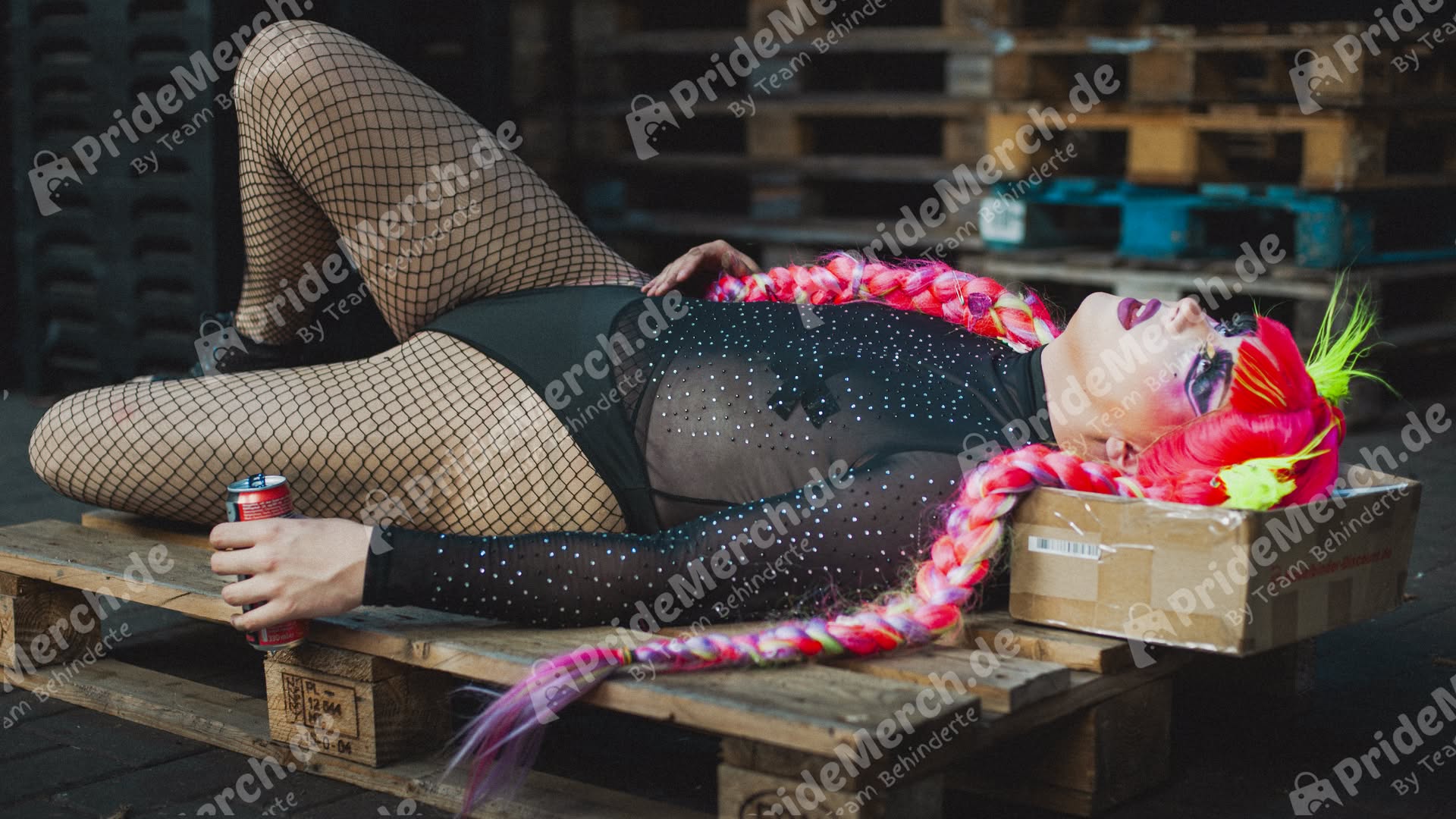 Drag Queen Luna Mattel liegt erschöpft im schwarzen Glitzerbody und mit neon-pinken Zöpfen auf einer rohen Holzpalette in einer industriellen Logistikhalle. Ihr Kopf ruht auf einem braunen Versandkarton. Ein politisches Fotomotiv, das Glamour und prekäre Arbeitsbedingungen kontrastiert.