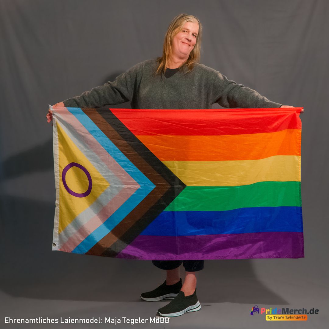 Laienmodell Maja Tegeler präsentiert die große Intersex Inclusive Progress Pride Flagge im Sitzen. Das farbintensive Polyestergewebe wird dabei dekorativ vor dem Oberkörper gehalten, während der Fokus auf den leuchtenden Community-Farben liegt.