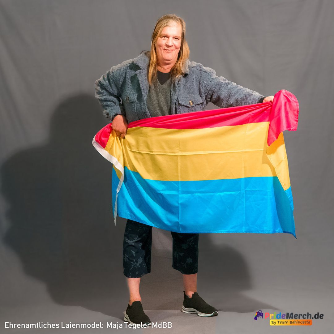 Das ehrenamtliche Laienmodell Maja Tegeler steht vor einem grauen Fotohintergrund. Sie hat langes blondes Haar und lächelt freundlich in die Kamera. Sie trägt eine dunkelgraue, flauschige Jacke, ein dunkles Oberteil, eine dunkelblaue Hose mit floralem Muster und schwarze Sneaker mit weißem Sohlenrand. Mit beiden Händen hält sie die große Pansexuell Pride Flagge auf Hüfthöhe locker vor sich gerafft, sodass die drei Streifen in Pink, Gelb und Blau weich nach unten fallen. Am linken Bildrand ist der weiße Saum