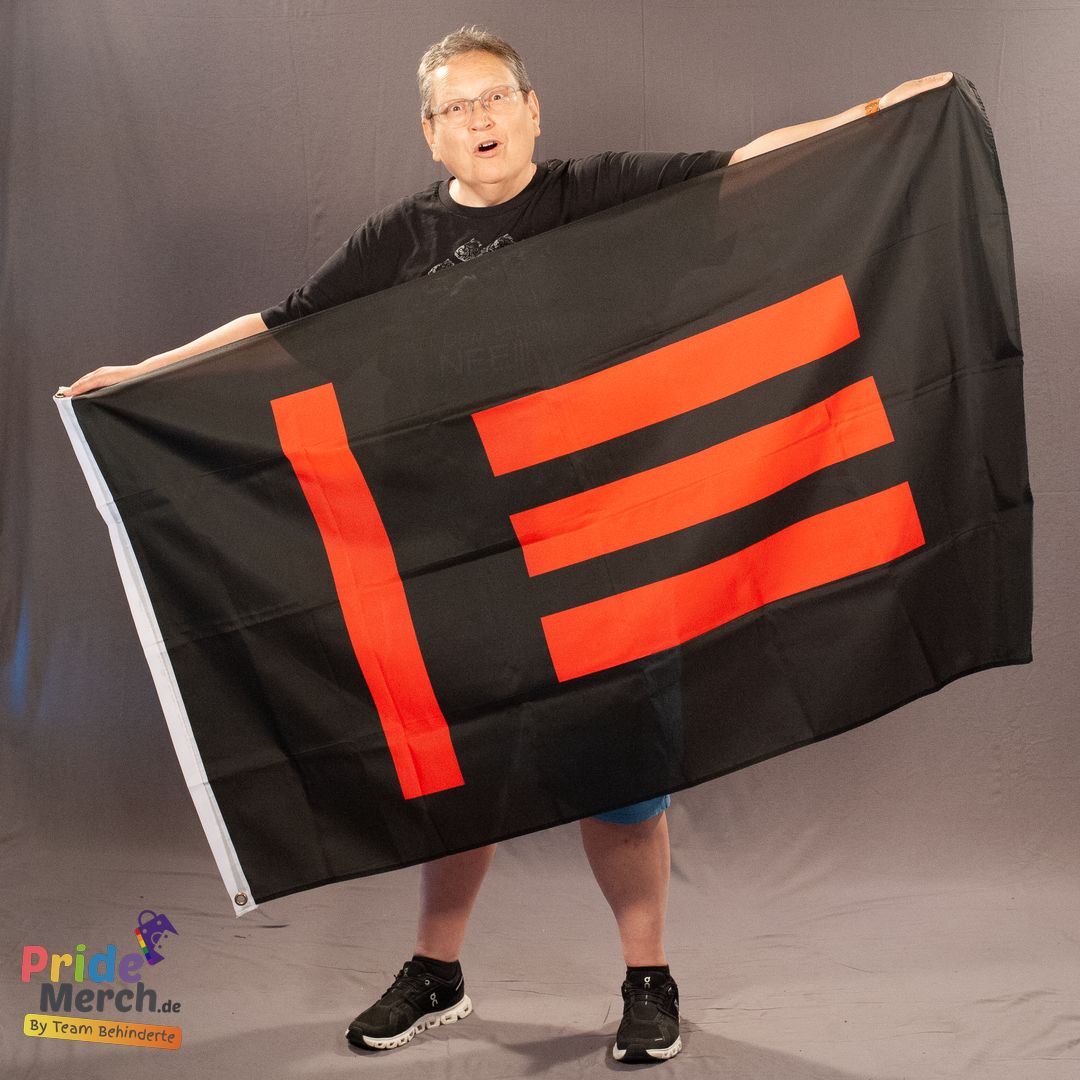 Das ehrenamtliche Laienmodell Beate Pellegrino steht vor einem grauen Hintergrund und hält die Master/Slave-Flagge mit beiden Händen weit aufgespannt vor sich. Beate trägt eine Brille, ein schwarzes T-Shirt, blaue Shorts und schwarze Sneaker. Auf der schwarzen Flagge leuchtet stark kontrastierend das rote M/s-Emblem: Eine vertikale Linie auf der linken Seite und drei horizontale Streifen, die nach rechts zeigen.