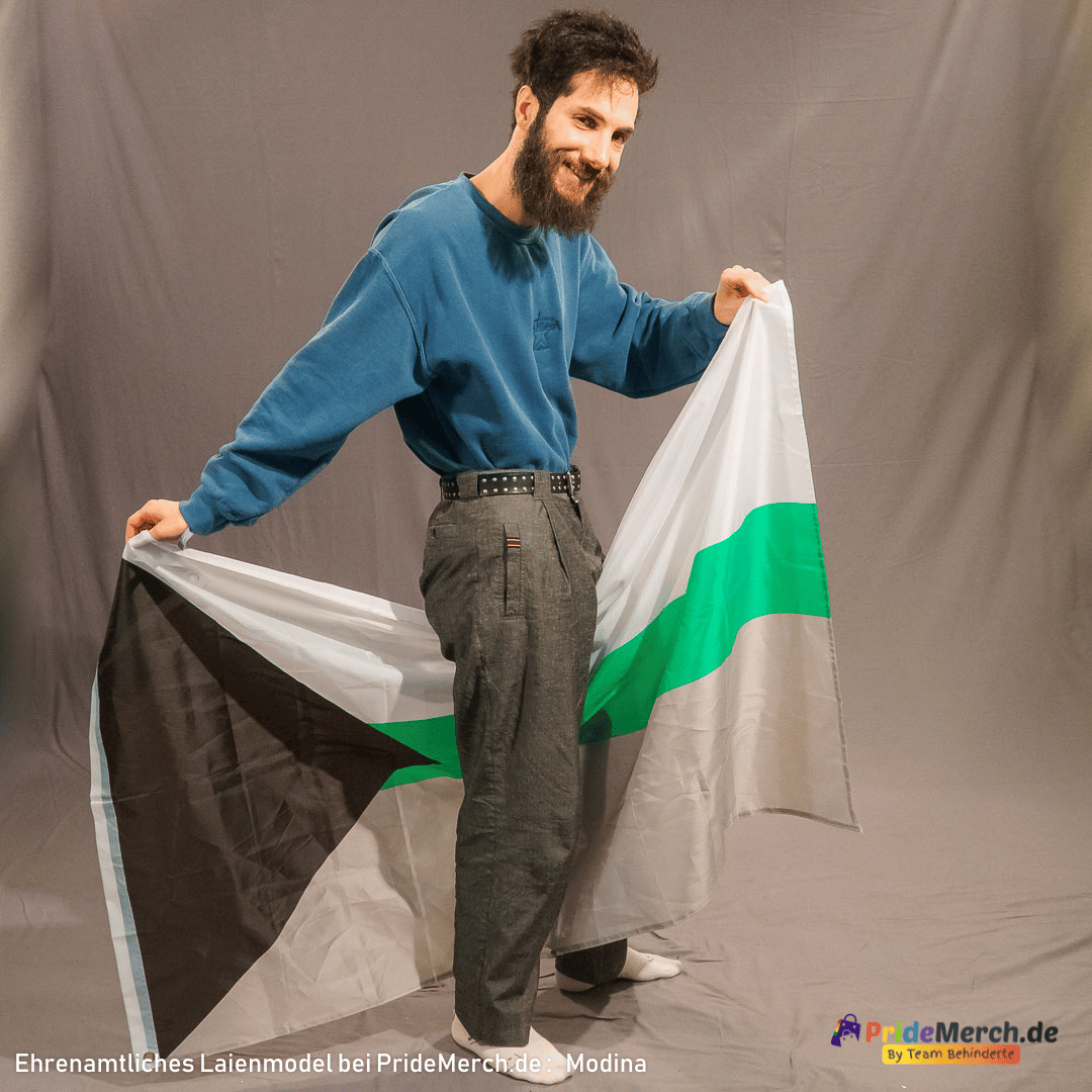 Modina, ein Mann mit dunklem Vollbart und dunklen Haaren, trägt einen blauen Pullover und eine dunkelgraue Hose. Er steht in einem Fotostudio und lächelt freundlich in die Kamera. Er hält die große demiromantische Flagge mit beiden Händen weit vor sich ausgebreitet. Die Flagge fällt in leichten Falten herab, wobei das schwarze Dreieck links und der grüne Mittelstreifen besonders gut zur Geltung kommen. Das Bild verdeutlicht die beeindruckende Größe der 150 cm breiten Flagge im Verhältnis zu einer Person.
