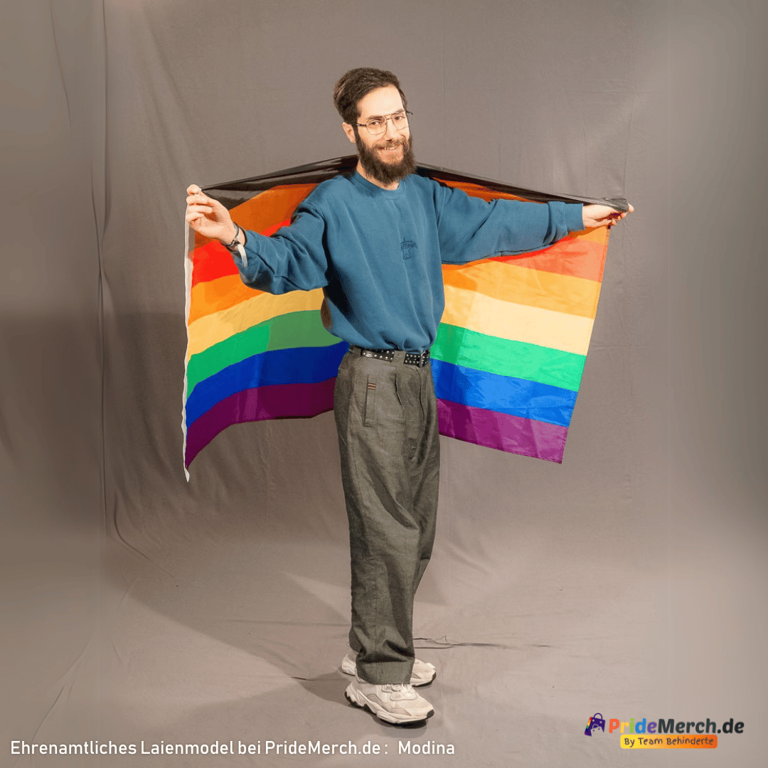 Das Laienmodell Modina von der Seite fotografiert trägt die inklusive Regenbogenflagge wie einen Umhang über den Schultern ausgebreitet. Die Aufhängungskante mit den Metallösen befindet sich auf der linken Seite.