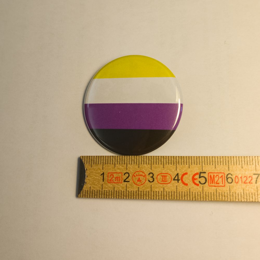Runder Metall-Button mit den vier waagerechten Streifen der Non-Binary-Flagge in Gelb, Weiß, Lila und Schwarz. Er liegt zur Veranschaulichung der Größe an einem hölzernen Zollstock, der etwa 37 Millimeter anzeigt.