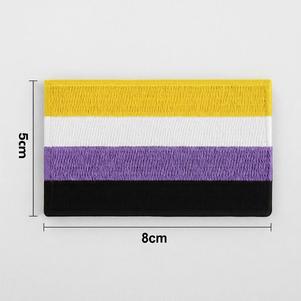 Ein gestickter Stoff-Aufnäher mit den vier horizontalen Streifen der Non-Binary Flagge in Gelb, Weiß, Violett und Schwarz. Der Patch hat eine sichtbare Textil-Struktur und liegt neben der Skala eines gelben Zollstocks, um die Maße für den Betrachter greifbar zu machen.