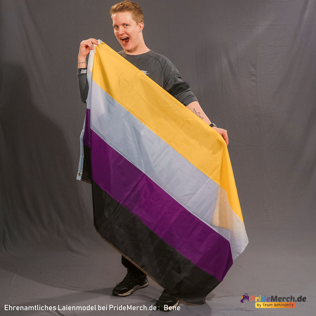 Bene lacht fröhlich mit geöffnetem Mund in die Kamera. Er rafft die große Non-Binary Flagge mit beiden Händen vor seinem Oberkörper zusammen, sodass die vier horizontalen Streifen in Gelb, Weiß, Lila und Schwarz in lockeren Falten fließen.
