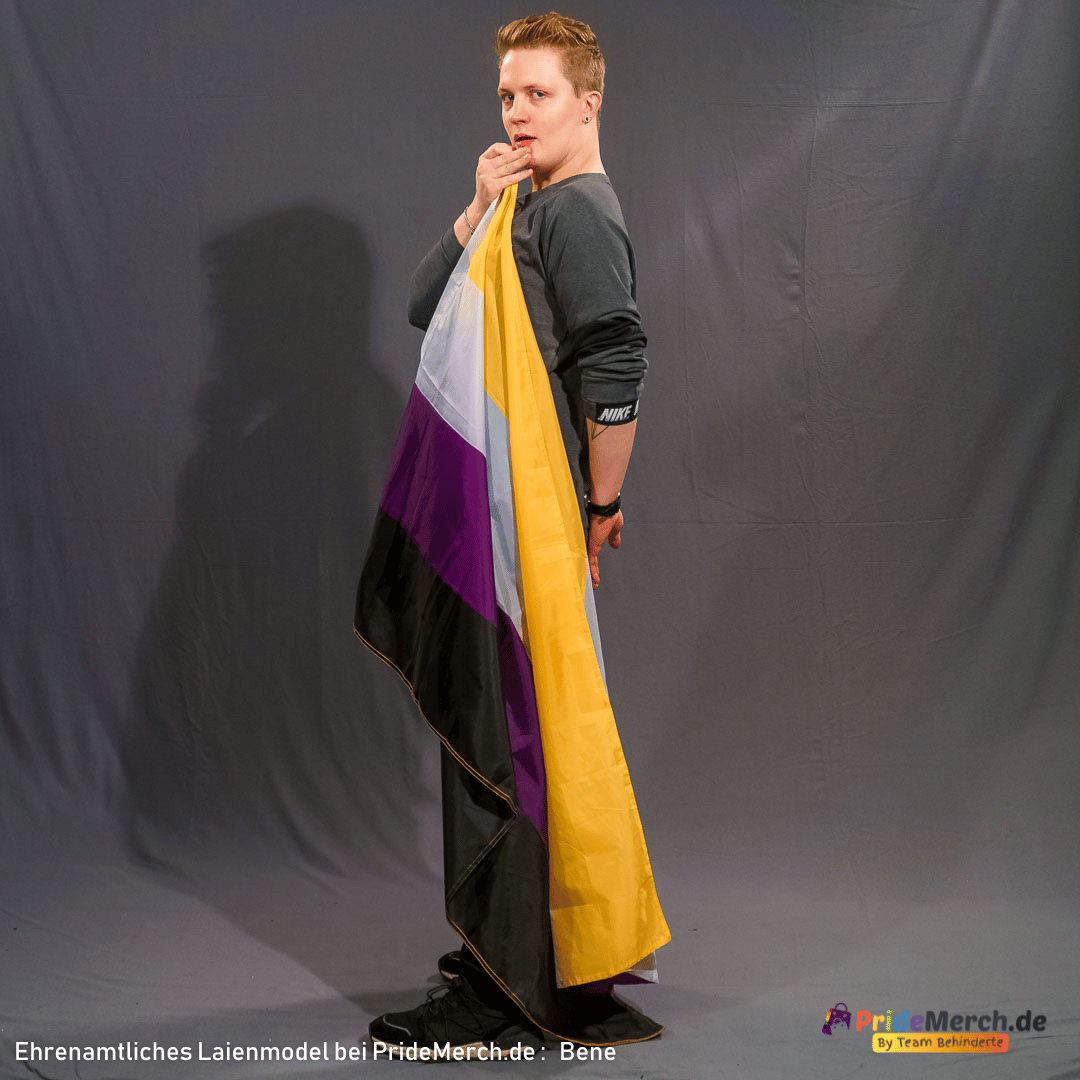 Das Laienmodell Bene steht seitlich vor einem grauen Hintergrund und blickt über seine linke Schulter intensiv in die Kamera. Er hat kurze, rötlich-blonde Haare und trägt ein dunkelgraues Langarmshirt. Die große Non-Binary Flagge mit ihren gelb, weiß, lila und schwarzen Streifen hat er sich wie ein schützendes Cape über den Rücken und die Schulter gelegt.