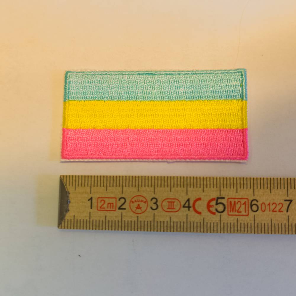 Ein rechteckiger Bügelaufnäher, dicht gestickt mit den drei horizontalen Farben der Pansexuell-Flagge: Pink, Gelb und Hellblau. Die glänzende Fadenstruktur ist gut sichtbar. Darunter liegt ein gelber Gliedermaßstab (Zollstock) zur genauen Größendarstellung.