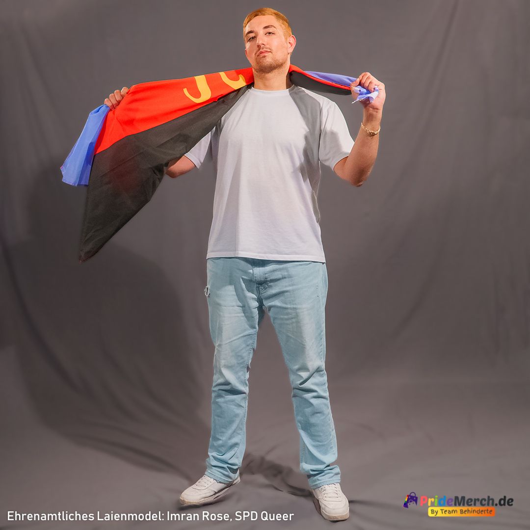 Das Laienmodell Imran Rose steht selbstbewusst vor einem grauen Hintergrund. Er hat kurze, kupferblonde Haare, trägt ein weißes T-Shirt und hellblaue Jeans. Die klassische Polyamorie Flagge hat er sich wie einen Umhang über den Rücken gelegt, wobei das gelbe Pi-Symbol über seiner rechten Schulter sichtbar ist.