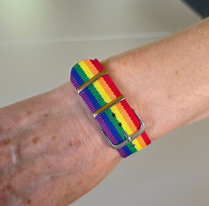 Das leuchtende Stoffarmband umschließt das Handgelenk einer Person. Aus dieser Perspektive sieht man auf die Innenseite des Handgelenks und erkennt, wie das Band mit seinen vertikalen Regenbogenstreifen eng und sicher auf der Haut anliegt. Eine schmale, polierte Metallschlaufe hält das doppelt liegende Nylonmaterial ordentlich zusammen.