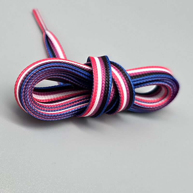 uf hellgrauem Untergrund liegt ein Paar flache Schnürsenkel, im Band abwechselnd in den fünf Farben der Genderfluid-Flagge (Pink, Weiß, Lila, Schwarz, Blau) gestreift und locker zu einer Schlaufe gelegt.