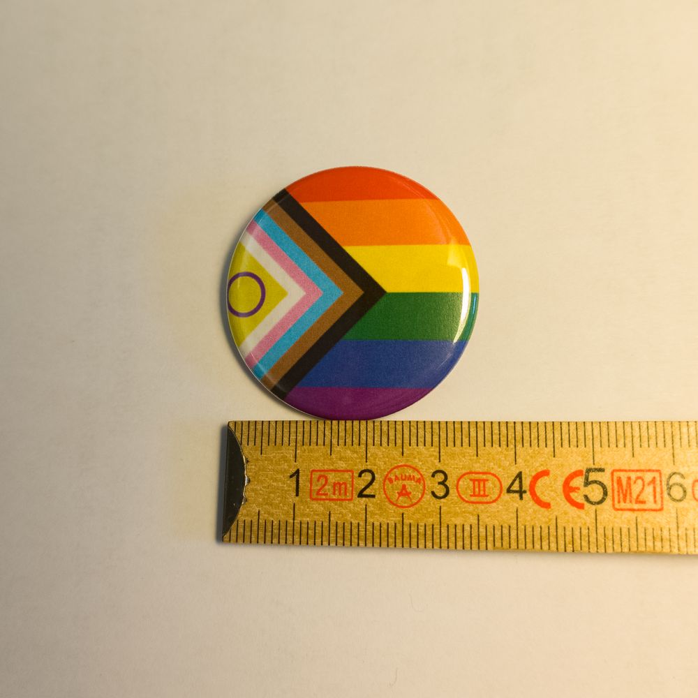 Runder Metall-Button mit dem Progress-Pride-Design. Neben dem Regenbogen sind die Pfeile sowie das gelbe Dreieck mit dem lila Kreis der Intersex-Community zu sehen. Der Pin liegt an einem Zollstock an und misst knapp 31 Millimeter.