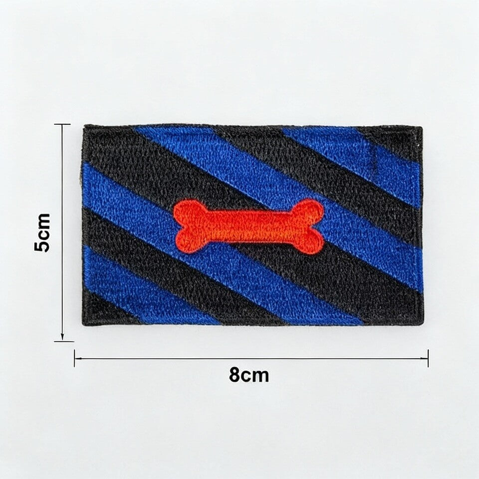 Ein textiler Aufnäher der Puppy Pride Flagge. Er zeigt abwechselnd blaue, schwarze und weiße diagonale Streifen, in der Mitte ist ein roter Hundeknochen gestickt. Der Aufnäher ist sauber umkettelt und liegt zur Größeneinordnung direkt neben einem gelben Zollstock.