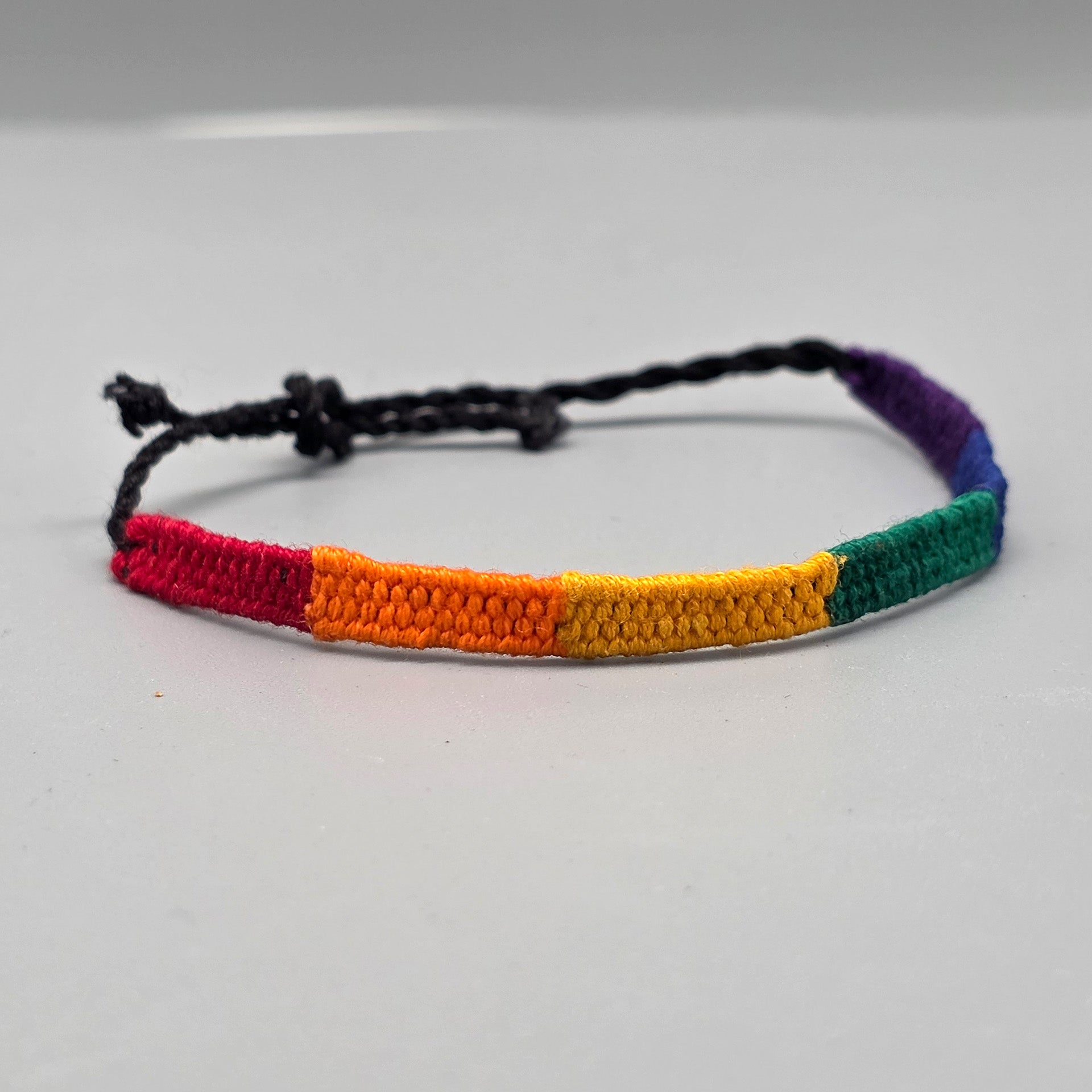 Das textile Regenbogen-Armband mit schwarzen Kordelenden ist von der linken Seite aus fotografiert. Die Nahaufnahme verdeutlicht den fließenden Übergang der dichten Webstruktur von sattem Rot über Orange und Gelb hin zu den kühleren Farbtönen.