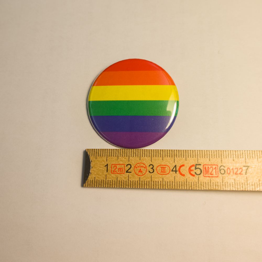 Runder Metall-Button mit den sechs Farben der klassischen Regenbogenflagge. Der glänzende Anstecker liegt zur Größeneinordnung neben einem ausgeklappten Zollstock und misst auf dem Foto etwa 37 Millimeter im Durchmesser.