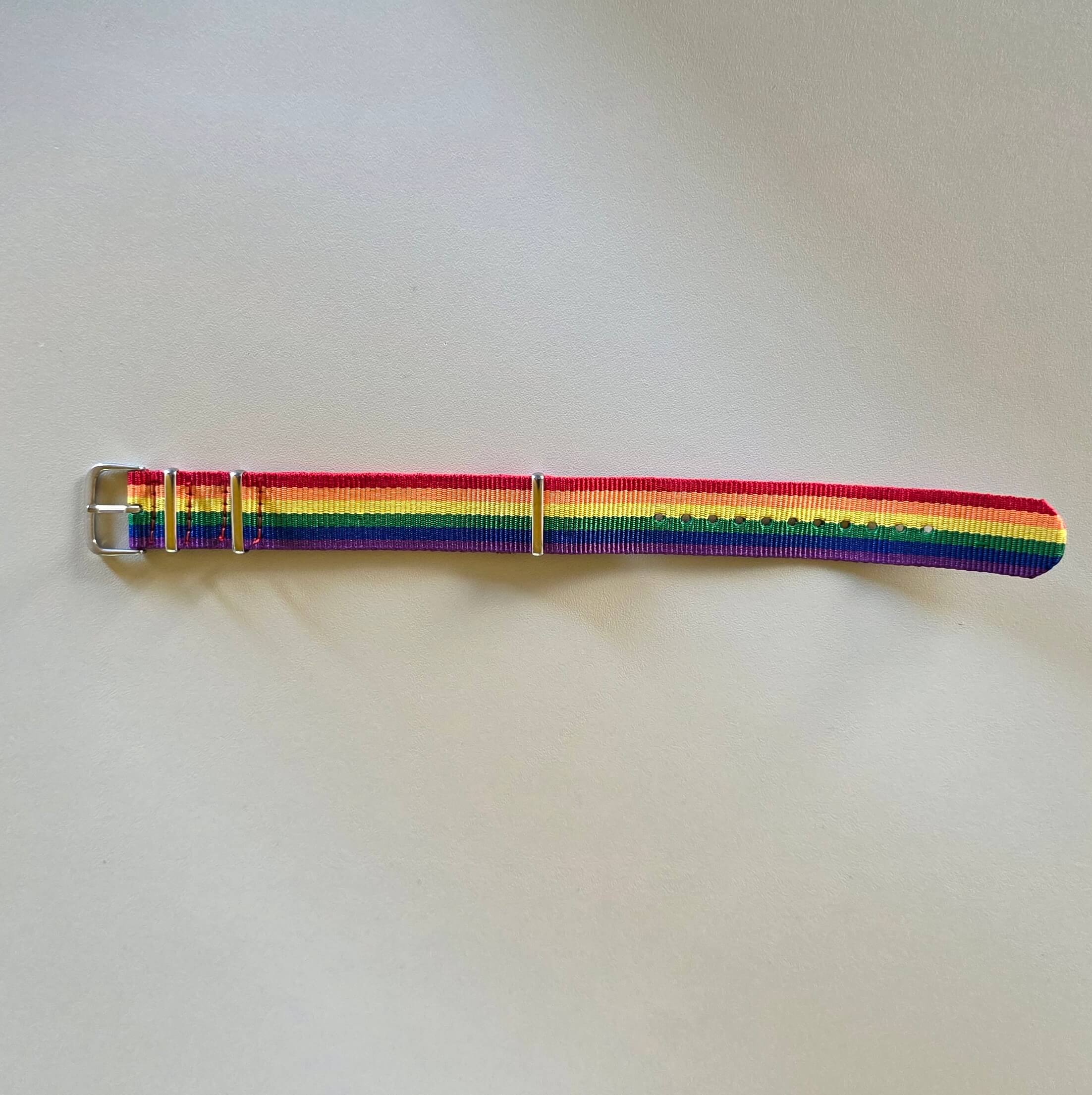 Ein flach ausgebreitetes, dicht gewebtes Nylonarmband liegt auf einem hellgrauen, glatten Untergrund. Die sechs Streifen der klassischen Pride-Flagge verlaufen längs und leuchten intensiv von Rot über Gelb bis Violett. Am linken Ende befindet sich eine silberfarbene Dornschnalle aus Metall, direkt gefolgt von zwei schmalen Metallschlaufen zur Fixierung.