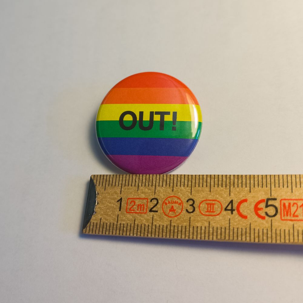 Glänzender, runder Metall-Button mit den sechs Farben der Regenbogenflagge. Mittig prangt der schwarze, ausdrucksstarke Schriftzug "OUT!". Zur Größeneinordnung liegt er neben einem Zollstock und zeigt 31 Millimeter Durchmesser.